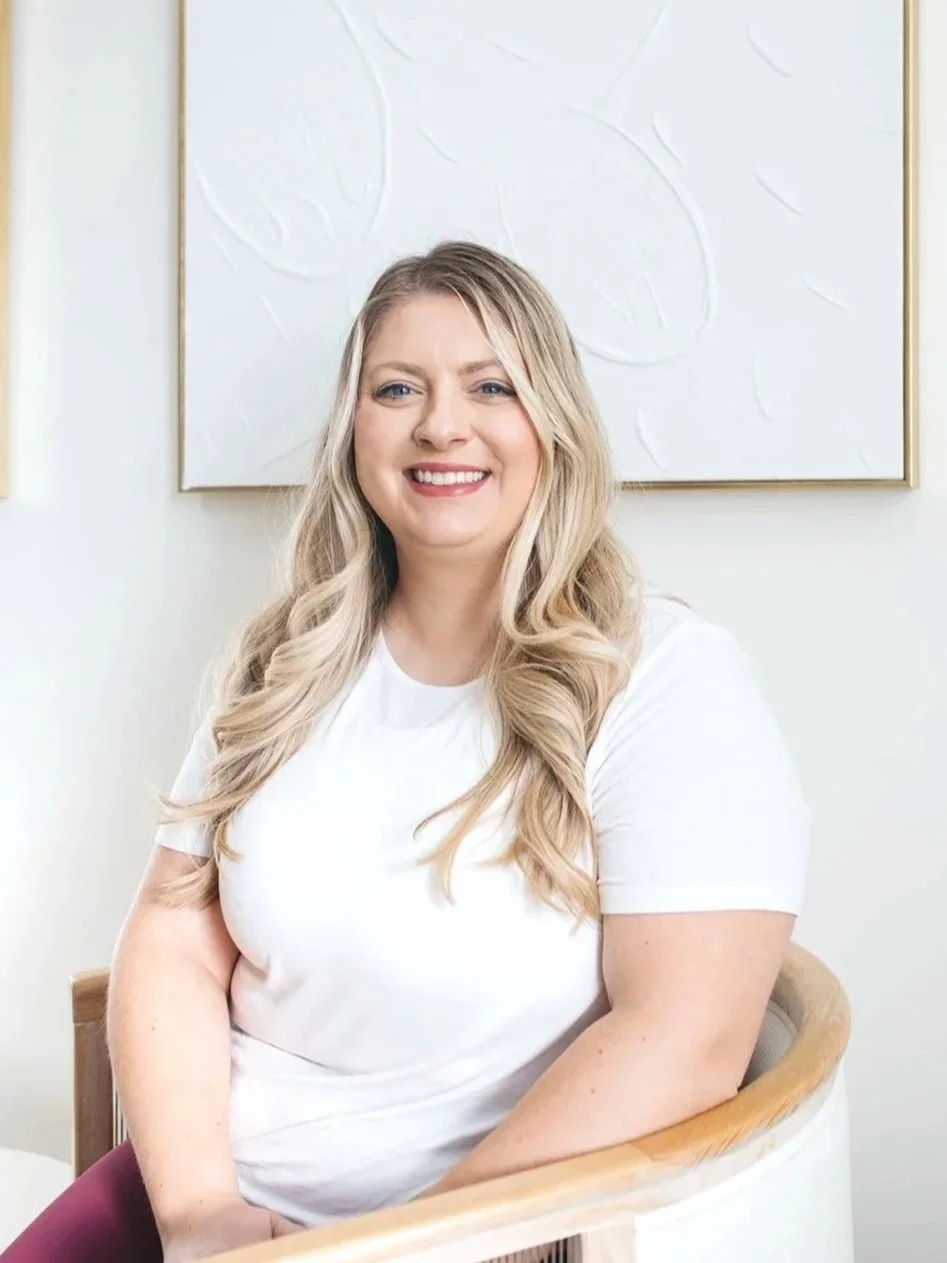 A woman with long blonde hair smiling and sitting on a wooden chair, wearing a white t-shirt, with abstract art on the wall behind her.