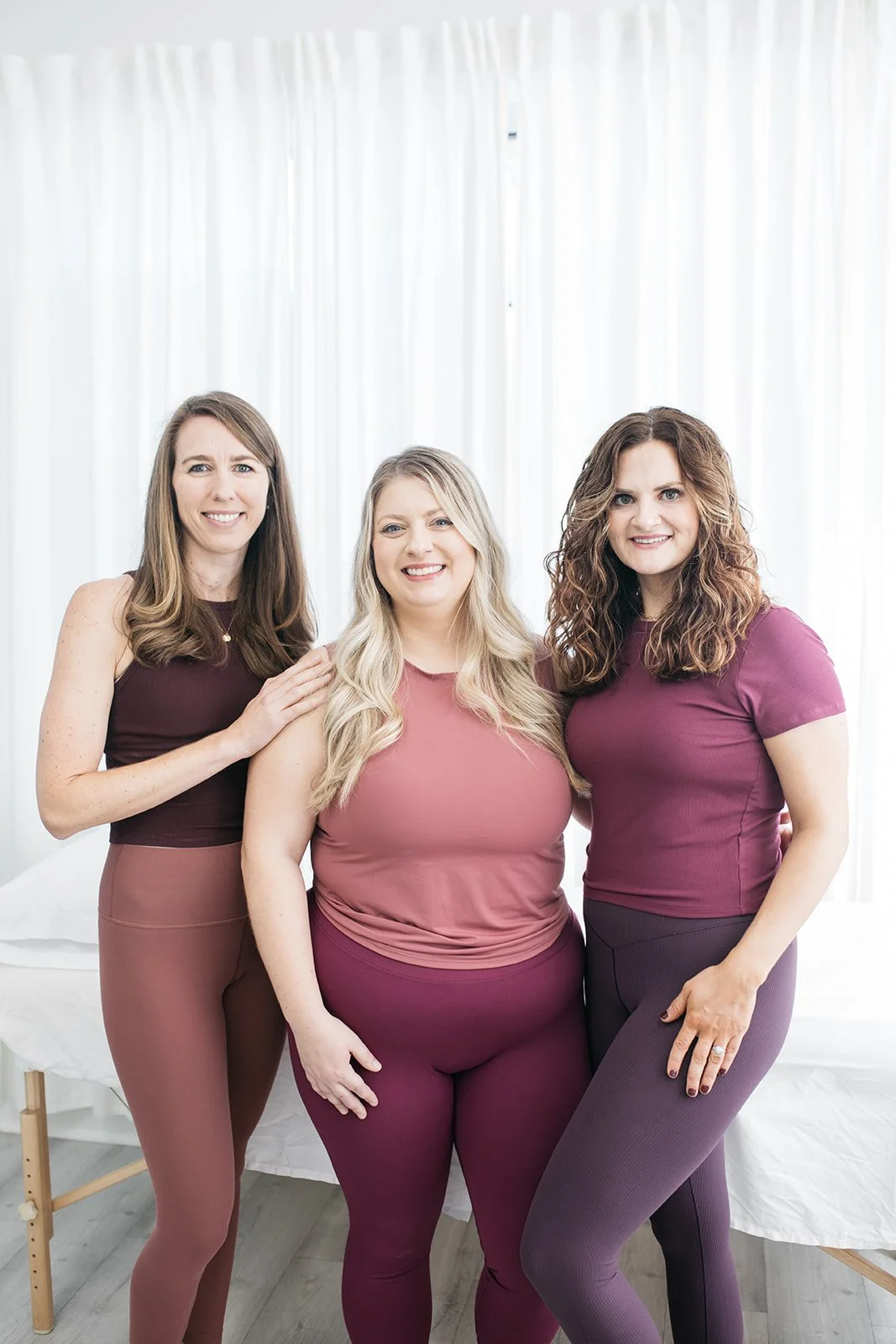 Three women smiling and standing together in front of white curtains, wearing matching maroon and pink outfits.