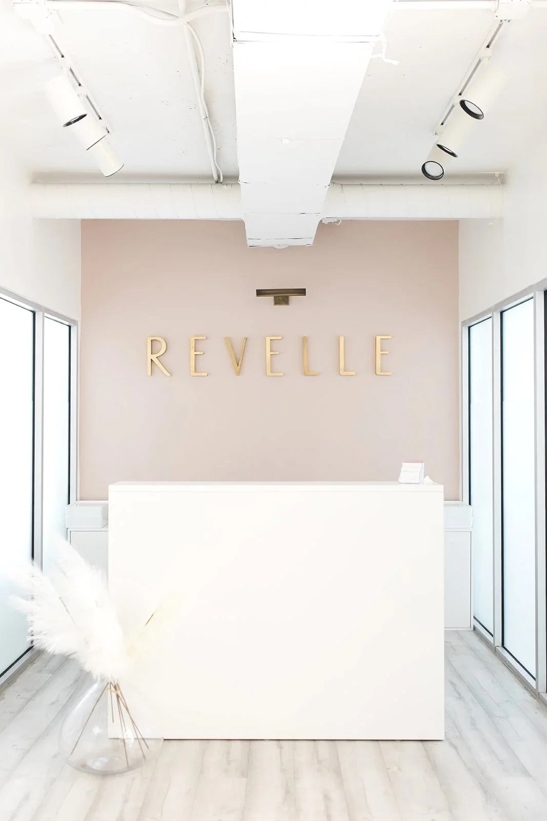 Minimalist reception area with a white desk, pastel pink wall with gold letters spelling "REVELLE," and large windows on both sides, bright with natural light.