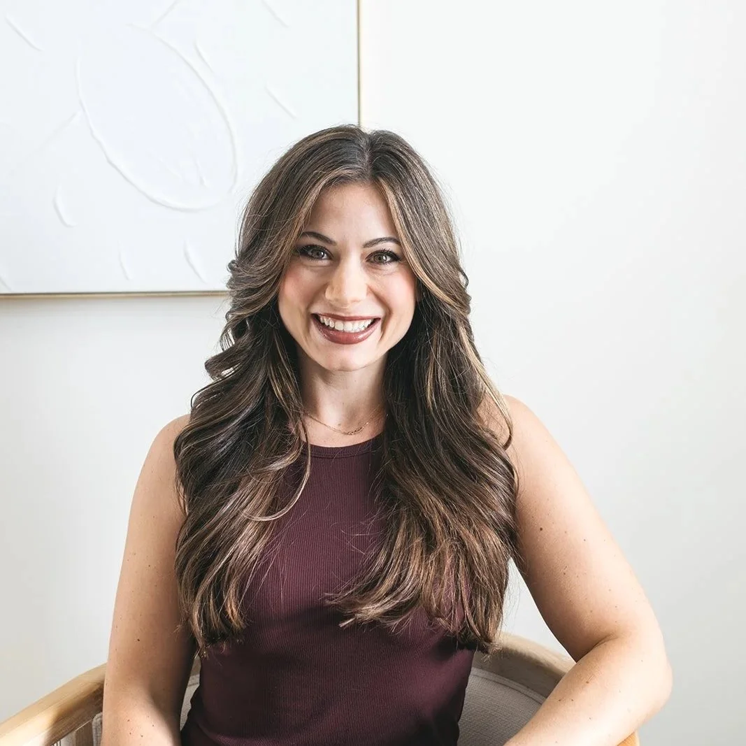A woman with long, wavy brown hair seated on a chair, smiling at the camera. She is wearing a white t-shirt, a watch, and denim jeans, with a light-colored background.