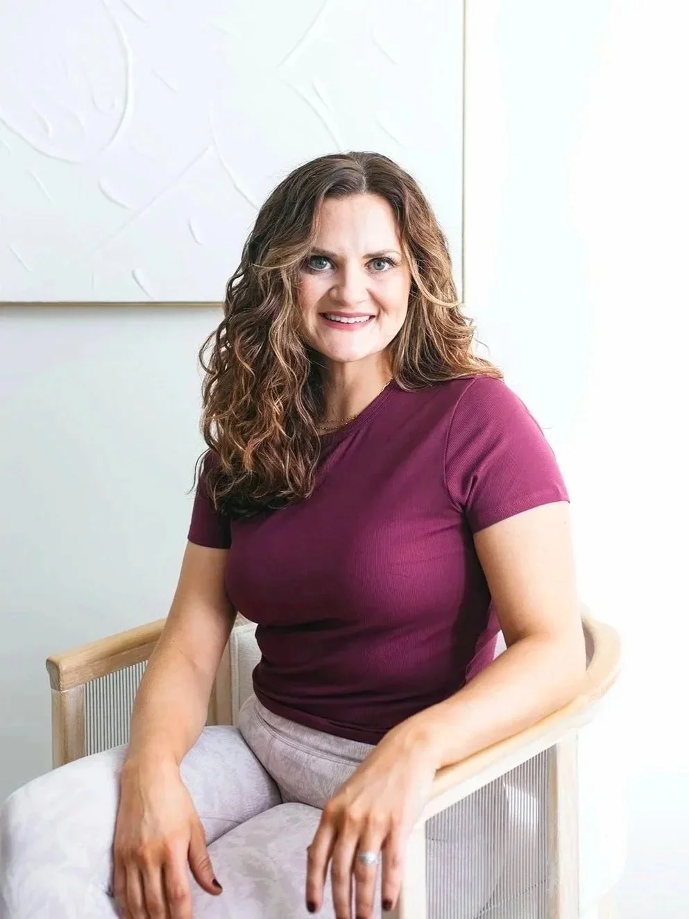 A smiling woman with brown curly hair wearing a burgundy t-shirt and light-colored pants, sitting on a wooden chair indoors.