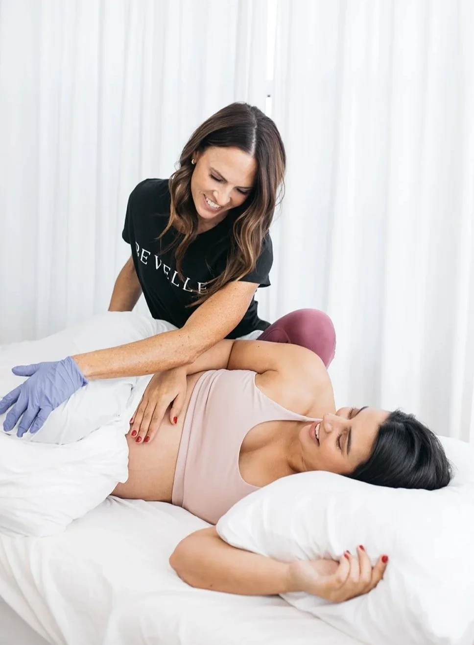 A woman with dark hair and a sleeveless beige top lying on a bed, smiling, as another woman with long wavy hair and a black shirt attends to her, wearing gloves, in a bright room with white curtains.