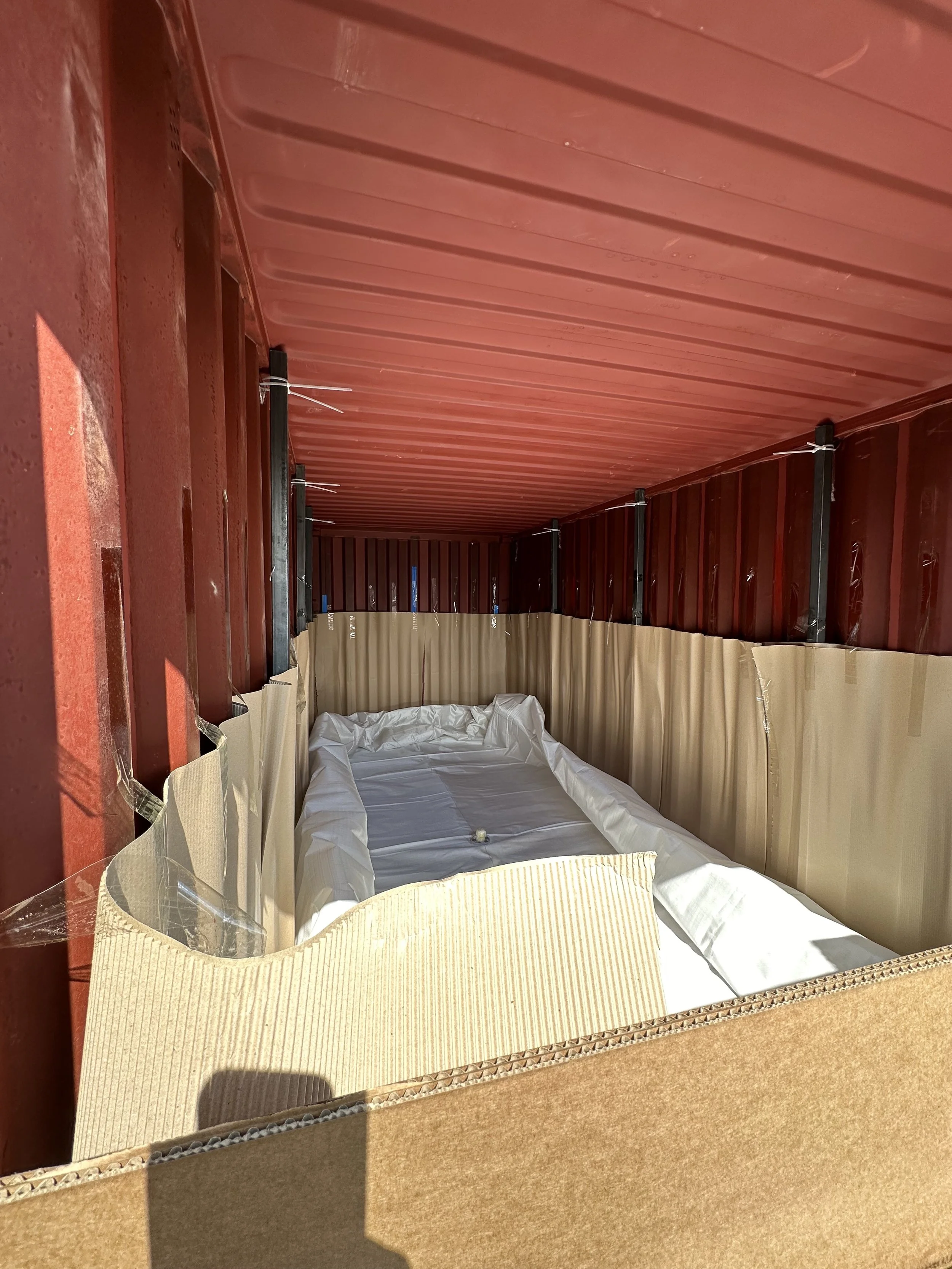 A red shipping container with an interior lined with cardboard and padding, containing a white mattress and foil-wrapped object.