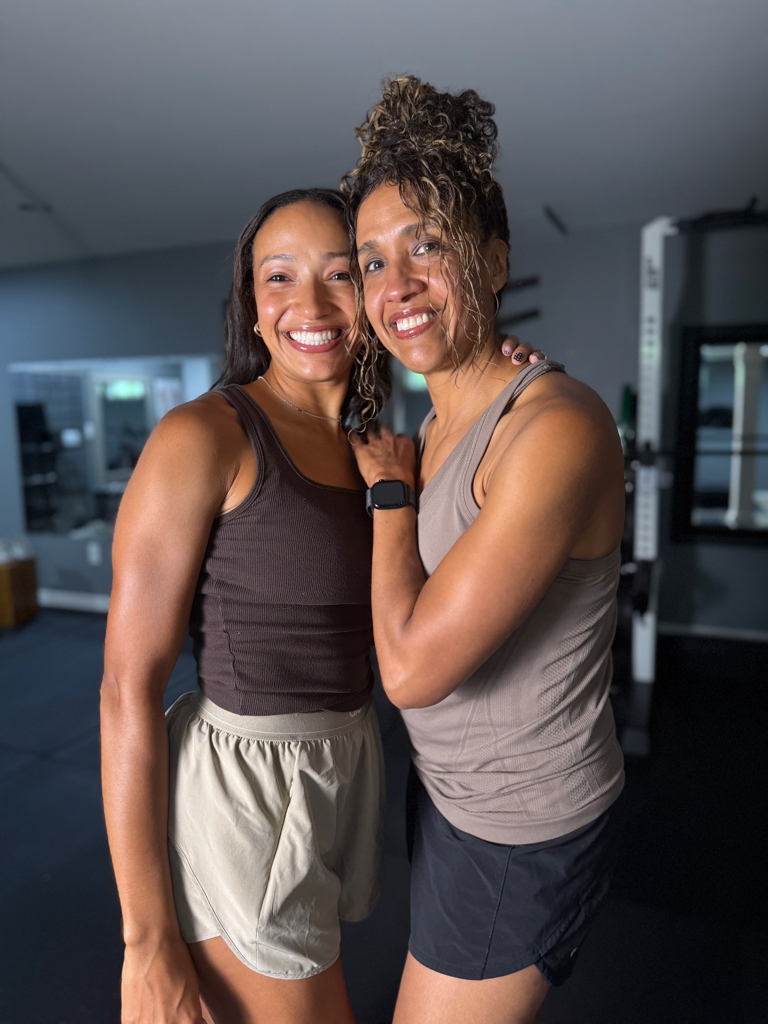 Two women smiling and hugging in a gym, dressed in workout clothes.