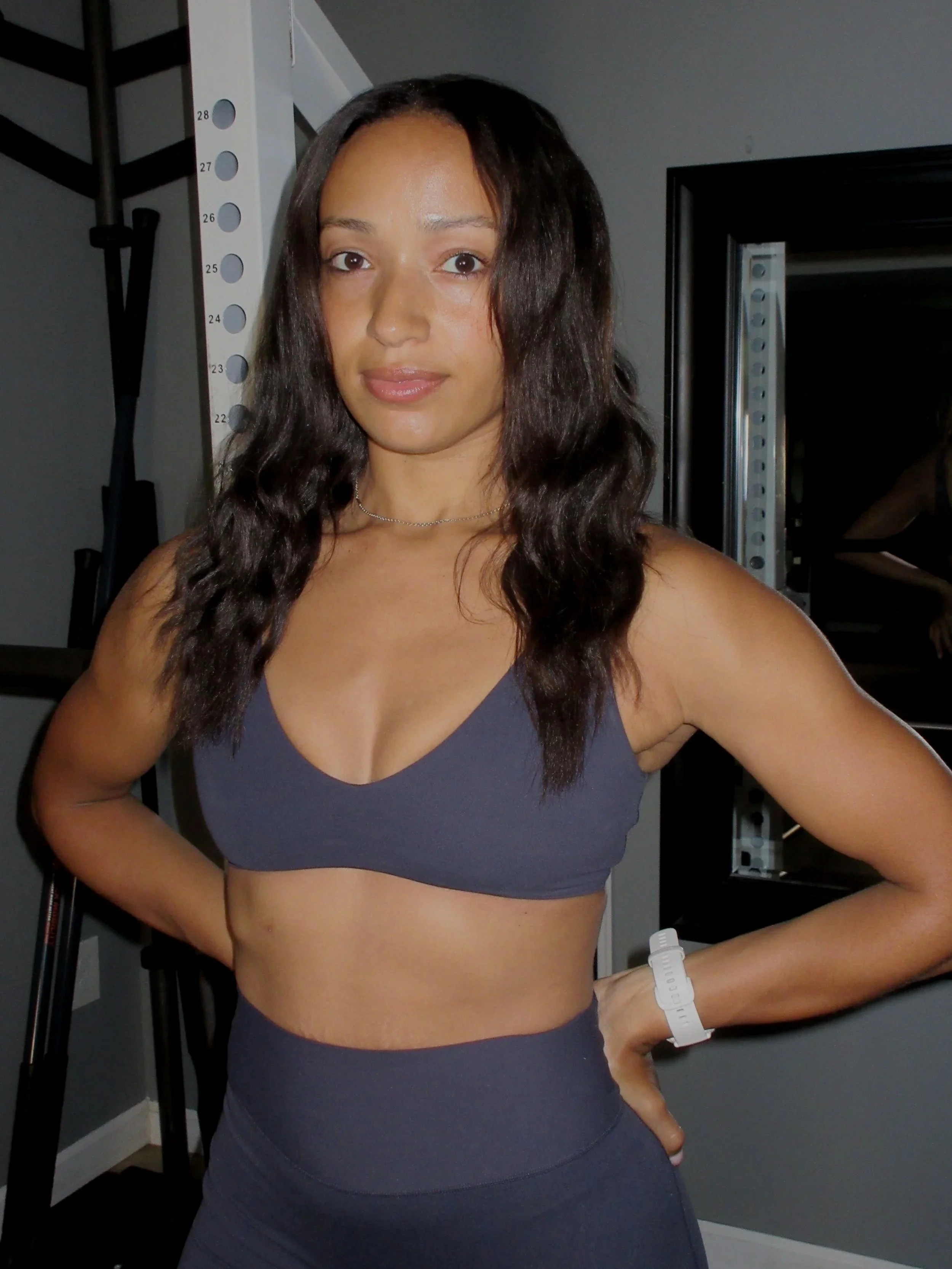 A woman standing in a gym, wearing a dark sports bra and matching workout pants, with one hand on her hip and a fitness tracker on her wrist, with gym equipment including a weight rack in the background.