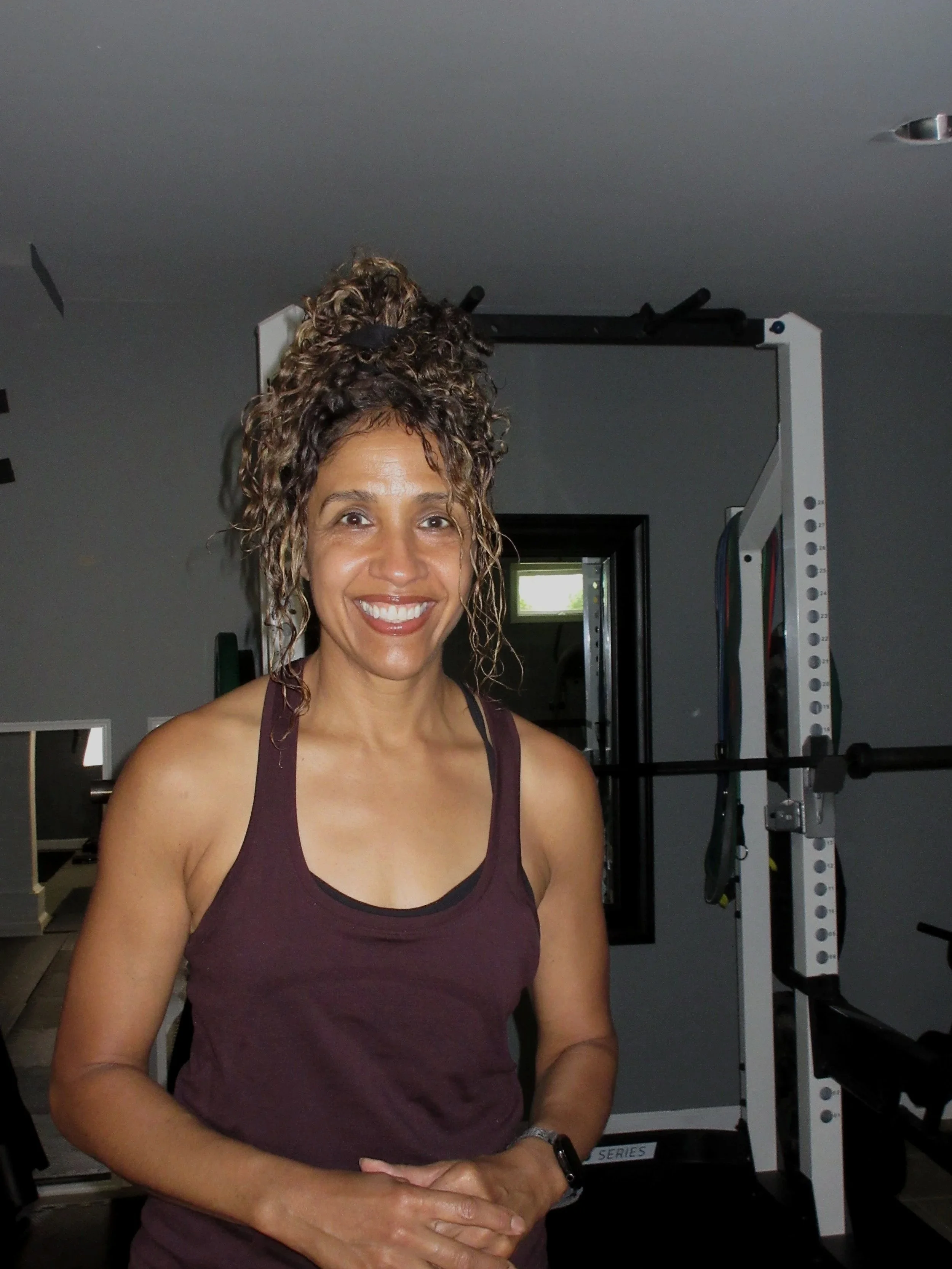 A woman with curly hair smiling, wearing a maroon tank top, standing in a gym with workout equipment in the background.