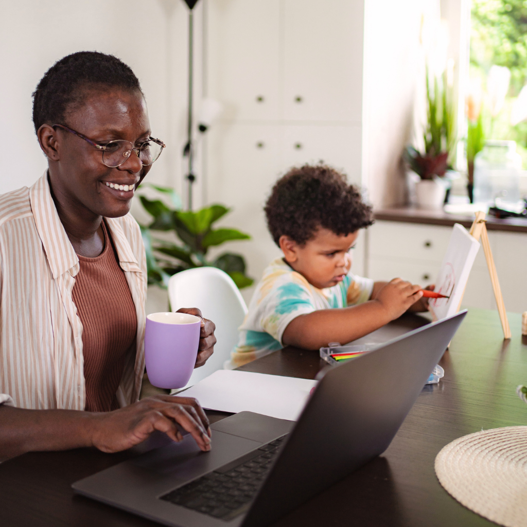 A woman and a young boy sitting at a wooden desk, with the woman helping the boy with a drawing tablet, keyboard, and mouse in front of them, in a bright room.