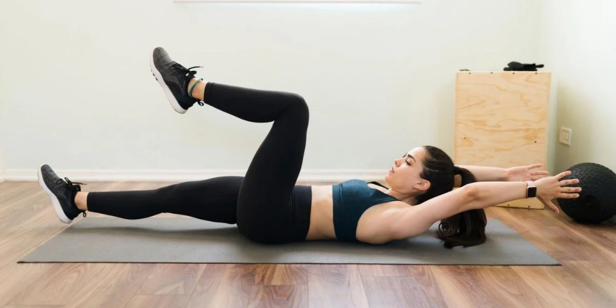 Woman performing the dead bug exercise on a gray mat in a home gym, demonstrating proper core engagement and spinal alignment