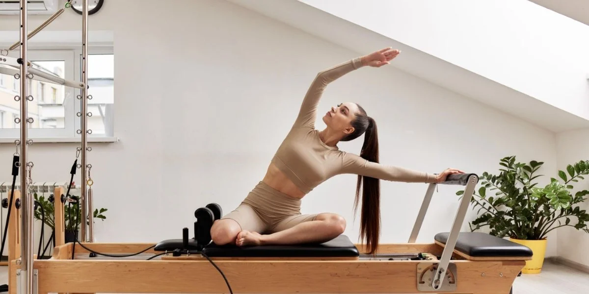 Workout plateau: a woman performs a seated side stretch on a Pilates reformer machine in a bright, plant-filled studio.