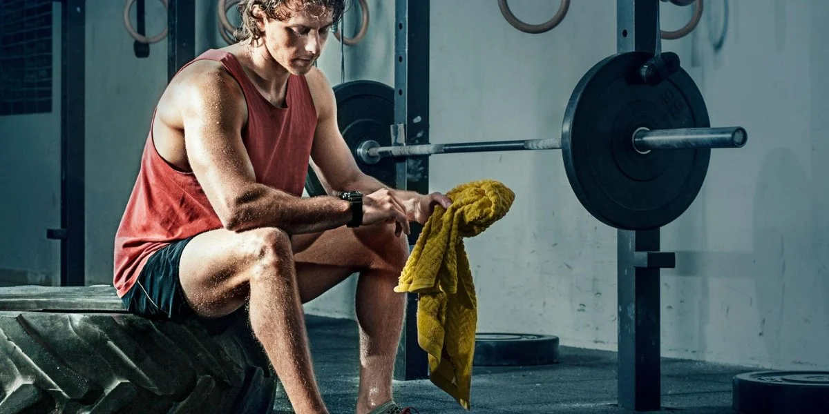 Progressive overload training male athlete resting on bench with barbell weights in gym environment showing strength training recovery and muscle development dedication.