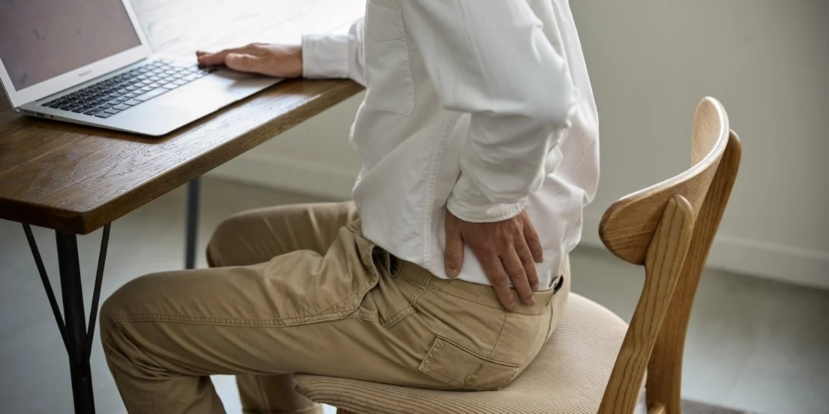 A desk worker sits at a table with a laptop while holding their lower back, highlighting the need for stretches for desk workers to ease sitting-related discomfort.