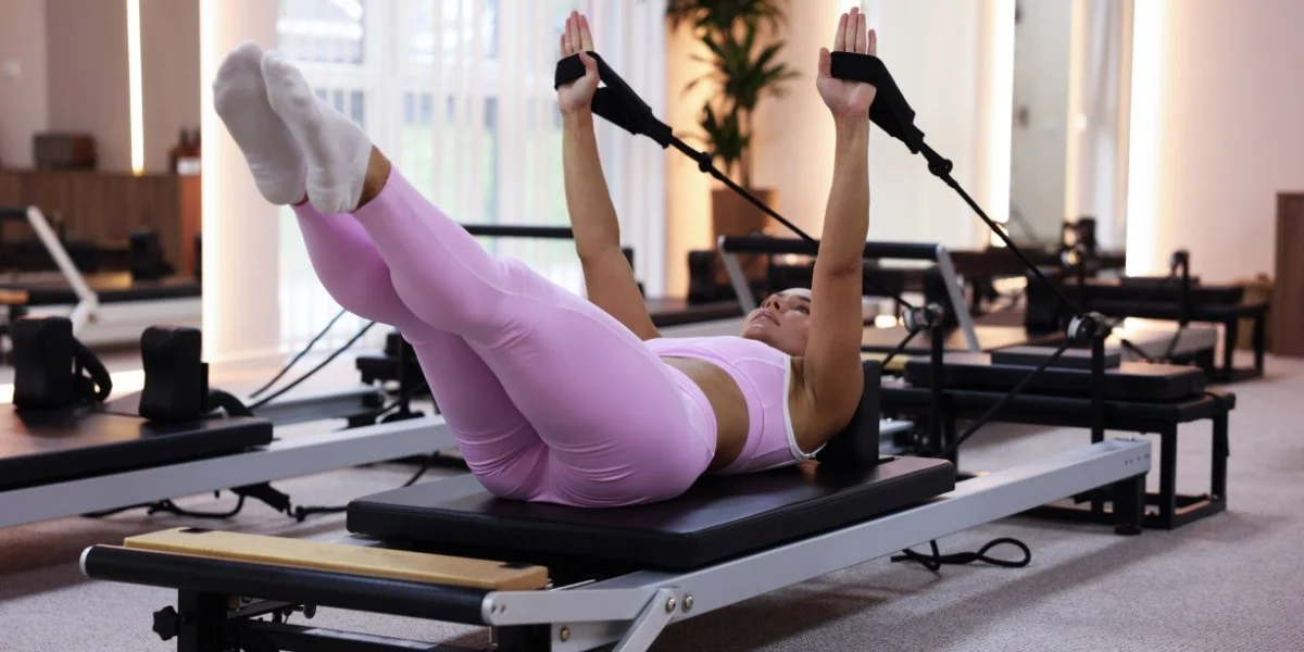 Bone strengthening activities - woman in pink athletic wear performing cable machine leg raises on a pilates reformer bed with resistance straps in a bright fitness studio with plants visible