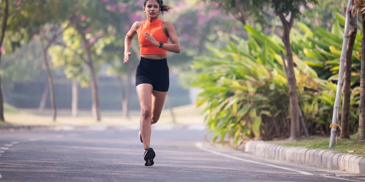 Bone strengthening activities - woman in orange sports top running outdoors on a tree-lined path wearing a fitness tracker, performing cardiovascular exercise in nature for bone health improvement