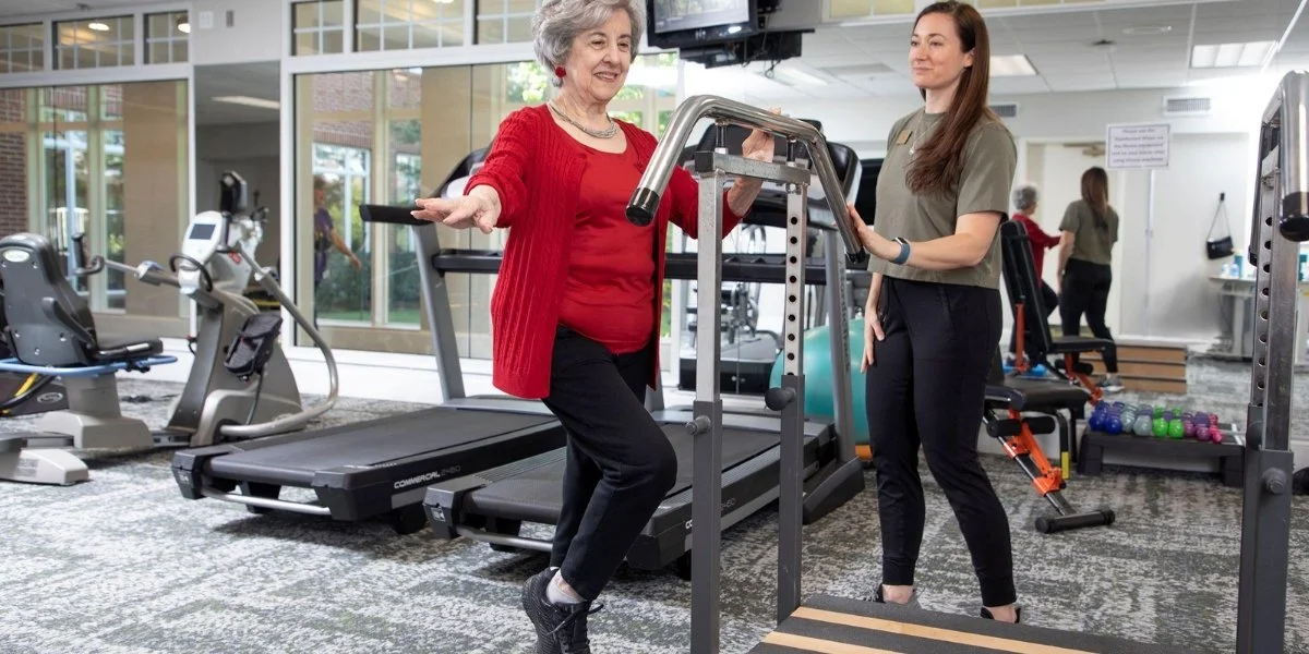 Balance training for seniors: an older woman uses an assisted trainer machine while a fitness professional guides her during a gym balance training workout.
