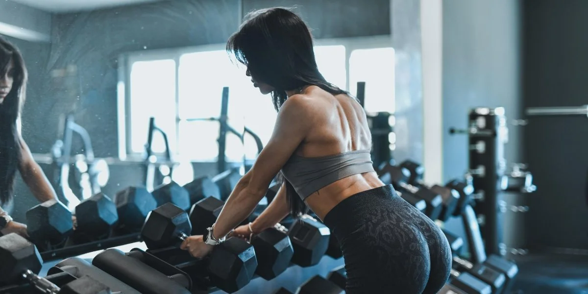 Woman lifting dumbbells in a modern gym environment, illustrating walking vs strength training for longevity through intensive weight training with mirrors and equipment visible in the background.