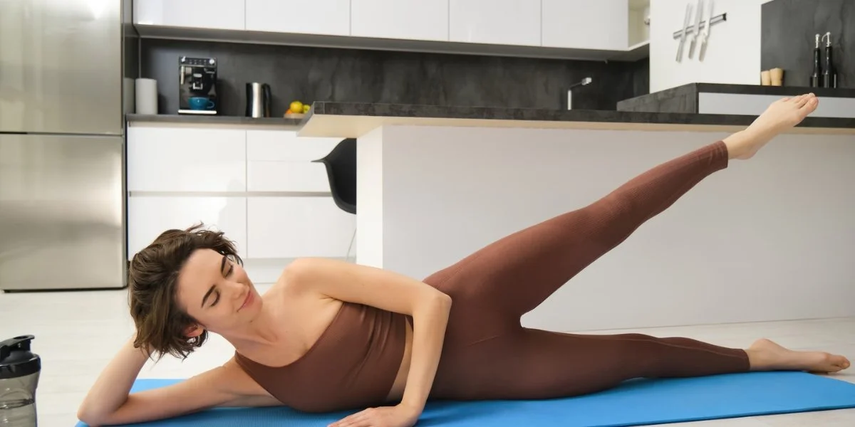 Side-lying leg raises exercise showing woman performing lateral leg lift on blue mat in modern kitchen setting, demonstrating hip and glute strengthening technique.
