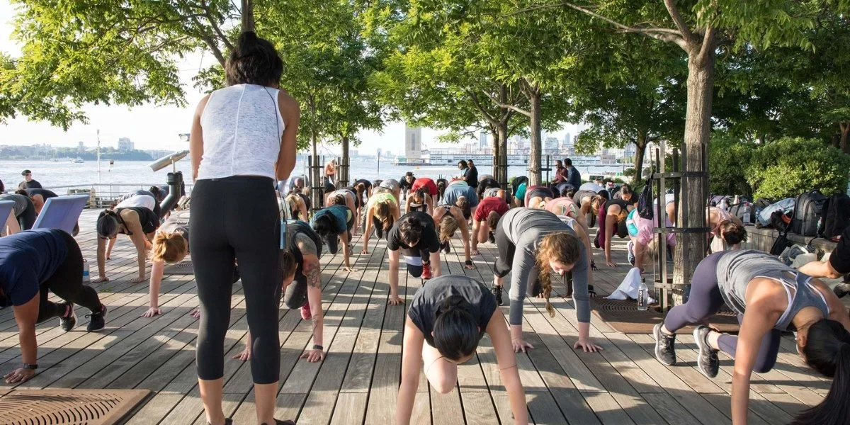 Large outdoor fitness group class at Chelsea Piers activities near the waterfront with participants exercising on a wooden deck overlooking the Hudson River and Manhattan skyline with tree coverage.