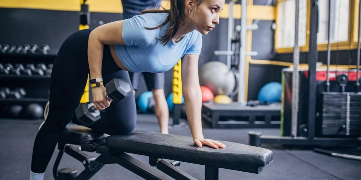 Woman performing dumbbell row exercise on weight bench demonstrating load used for the same exercise over time progression in modern gym with fitness equipment