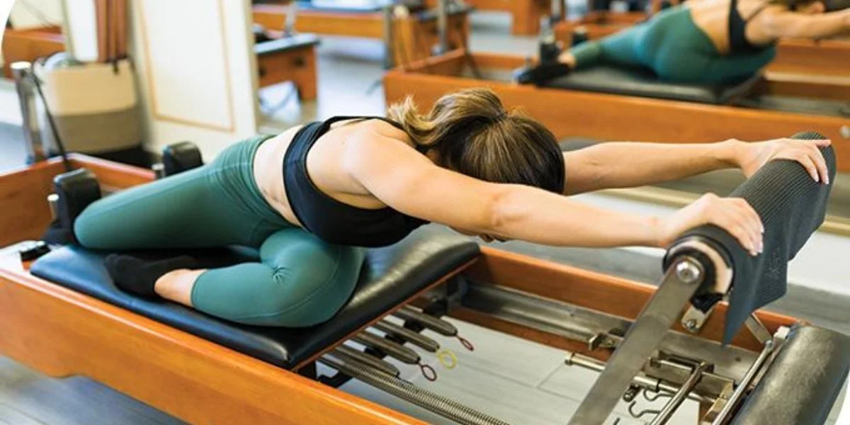 A woman stretches forward in a resting pose on a pilates reformer, illustrating the calm, sustainable approach behind consistency over intensity.