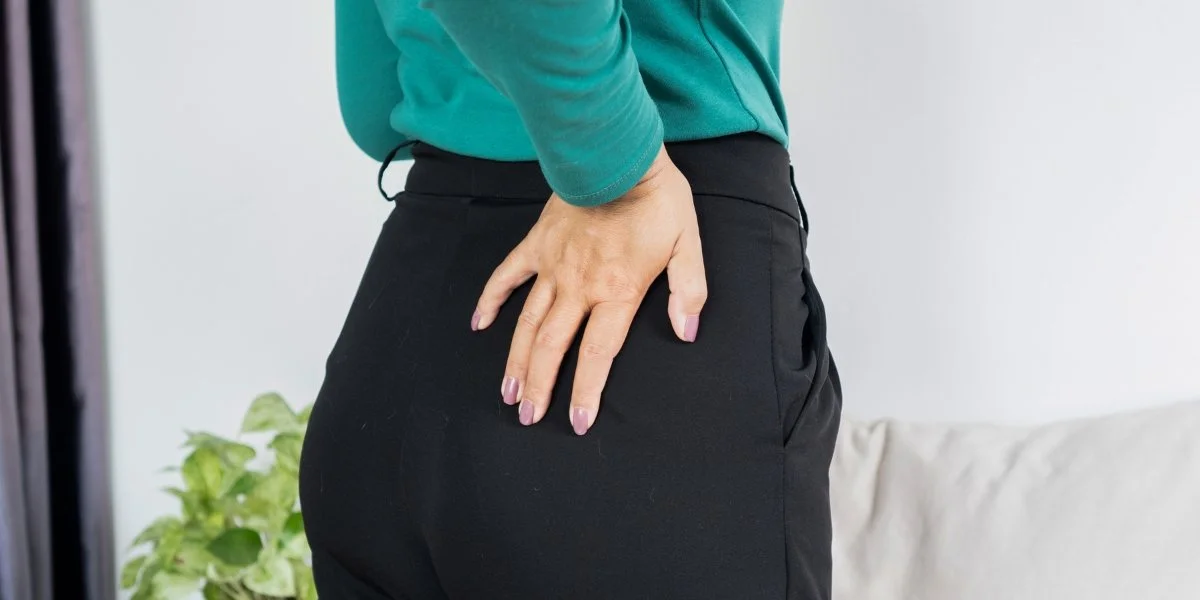 Why are my hips so tight - person in teal sweater placing hands on lower back and hip area while wearing black pants, demonstrating hip tightness and discomfort in a home setting with plants visible