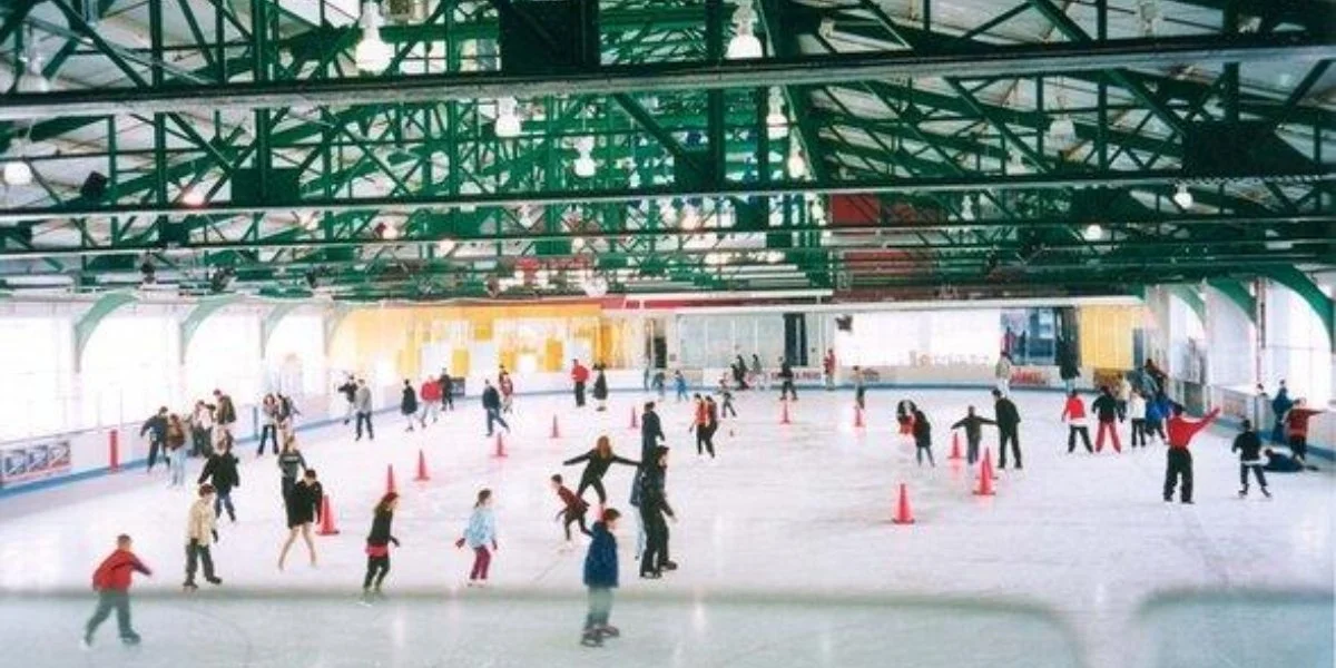 Indoor ice skating rink at Chelsea Piers activities featuring Sky Rink with numerous skaters of various ages enjoying the sport under a glass roof with exposed green steel beam structure.