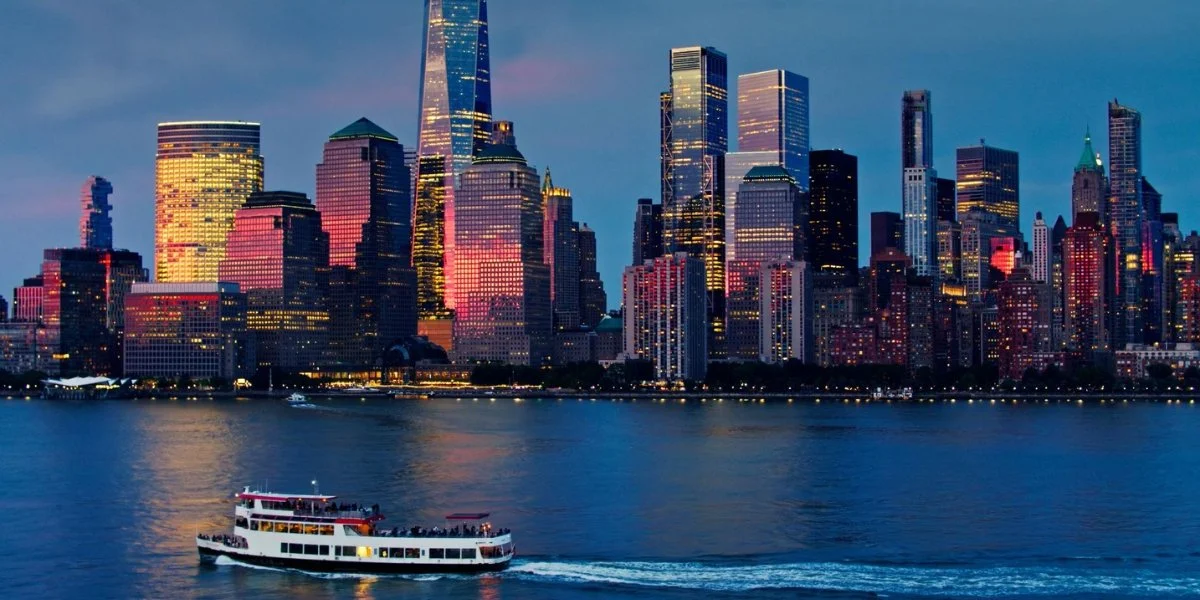 Scenic harbor tour boat sailing on the Hudson River with illuminated Manhattan skyline featuring iconic skyscrapers at dusk, representing Chelsea Piers activities including waterfront sightseeing and harbor cruises.