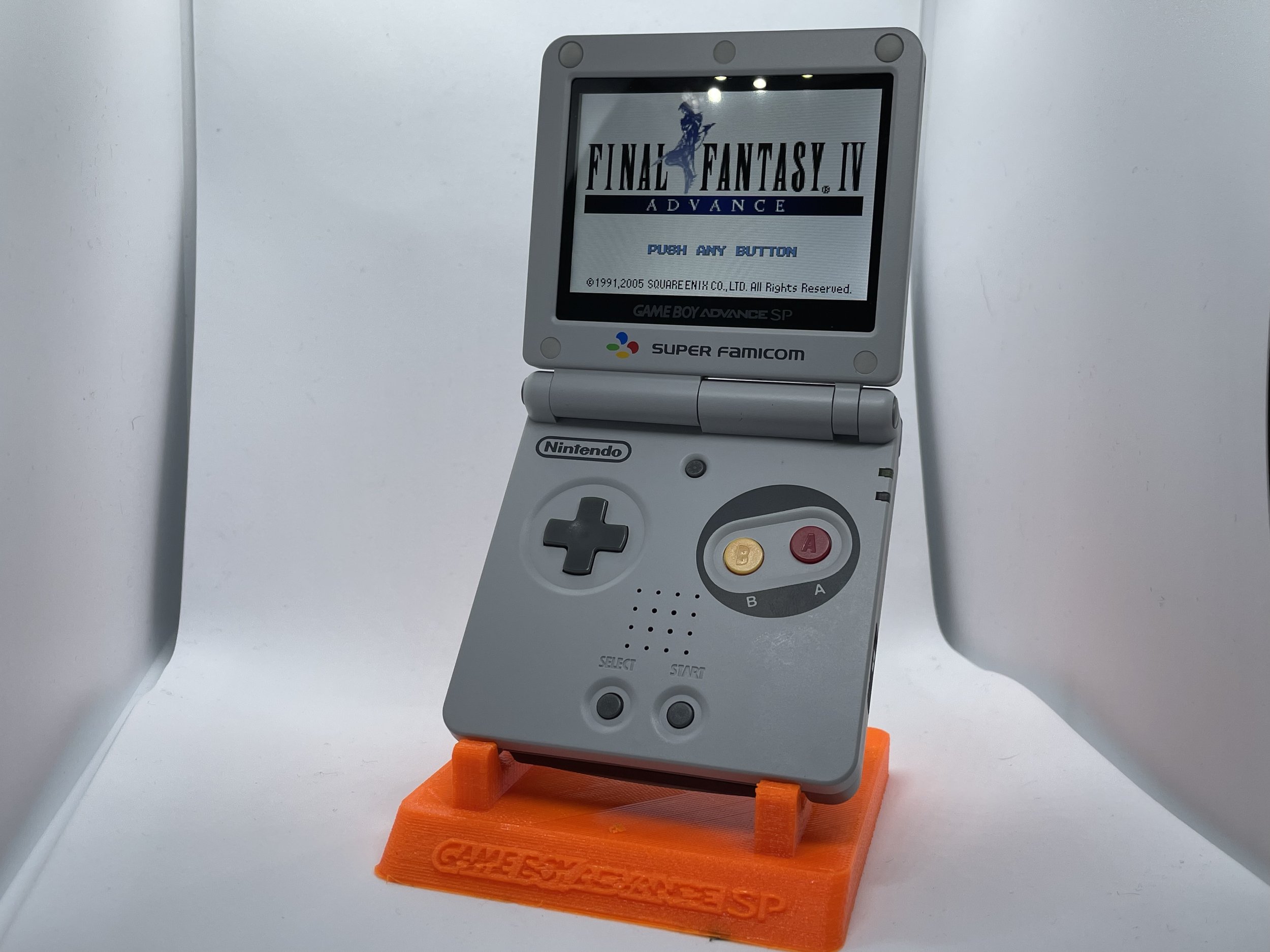 A gray Nintendo Game Boy Advance SP with a screen displaying the title screen of Final Fantasy IV Advance. The device is placed on an orange stand with Game Explosion written on it.
