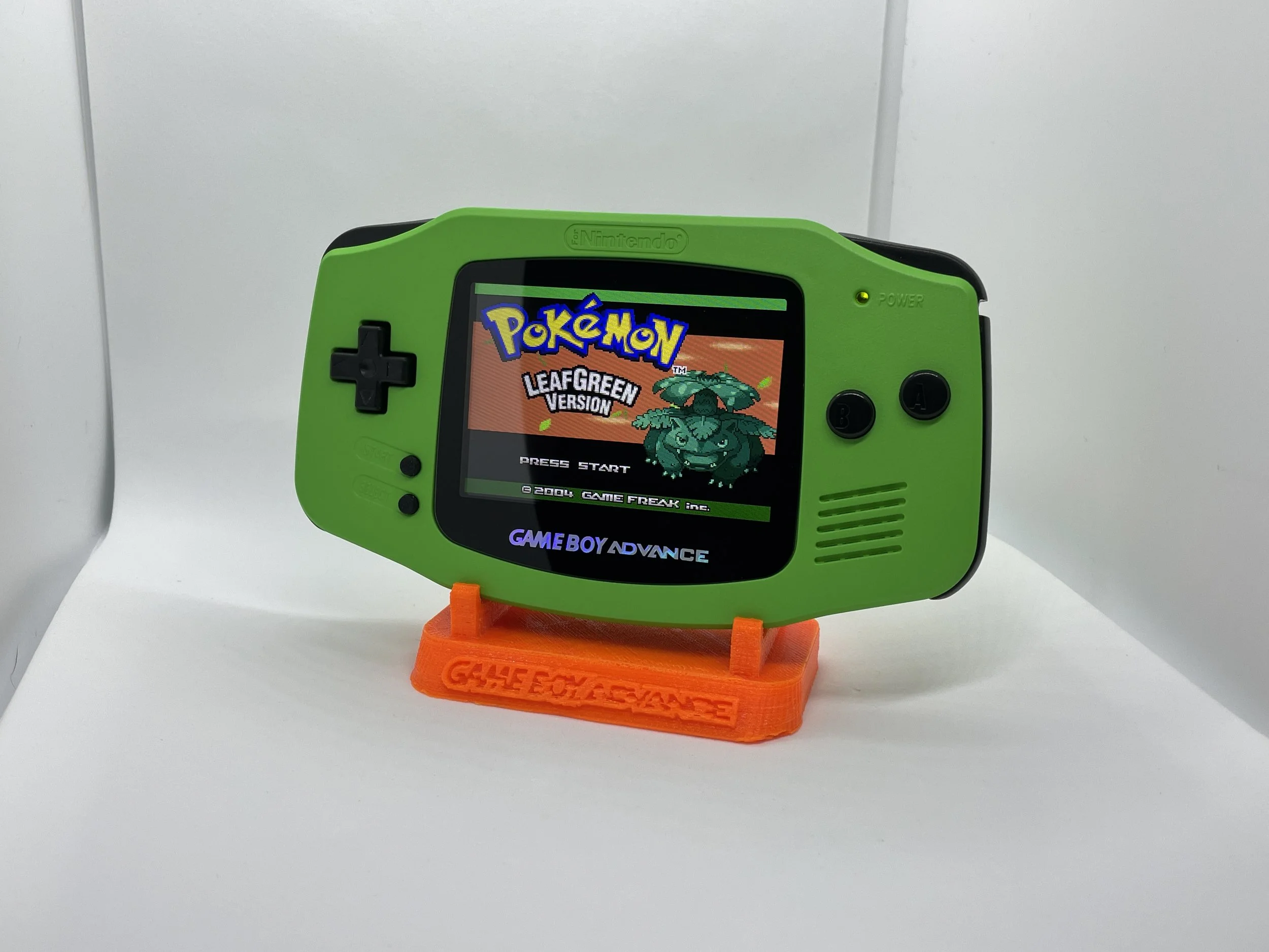 Green Nintendo Game Boy Advance showing Pokémon LeafGreen Version on the screen, placed on an orange stand with 'GAME BOY ADVANCE' written on it.