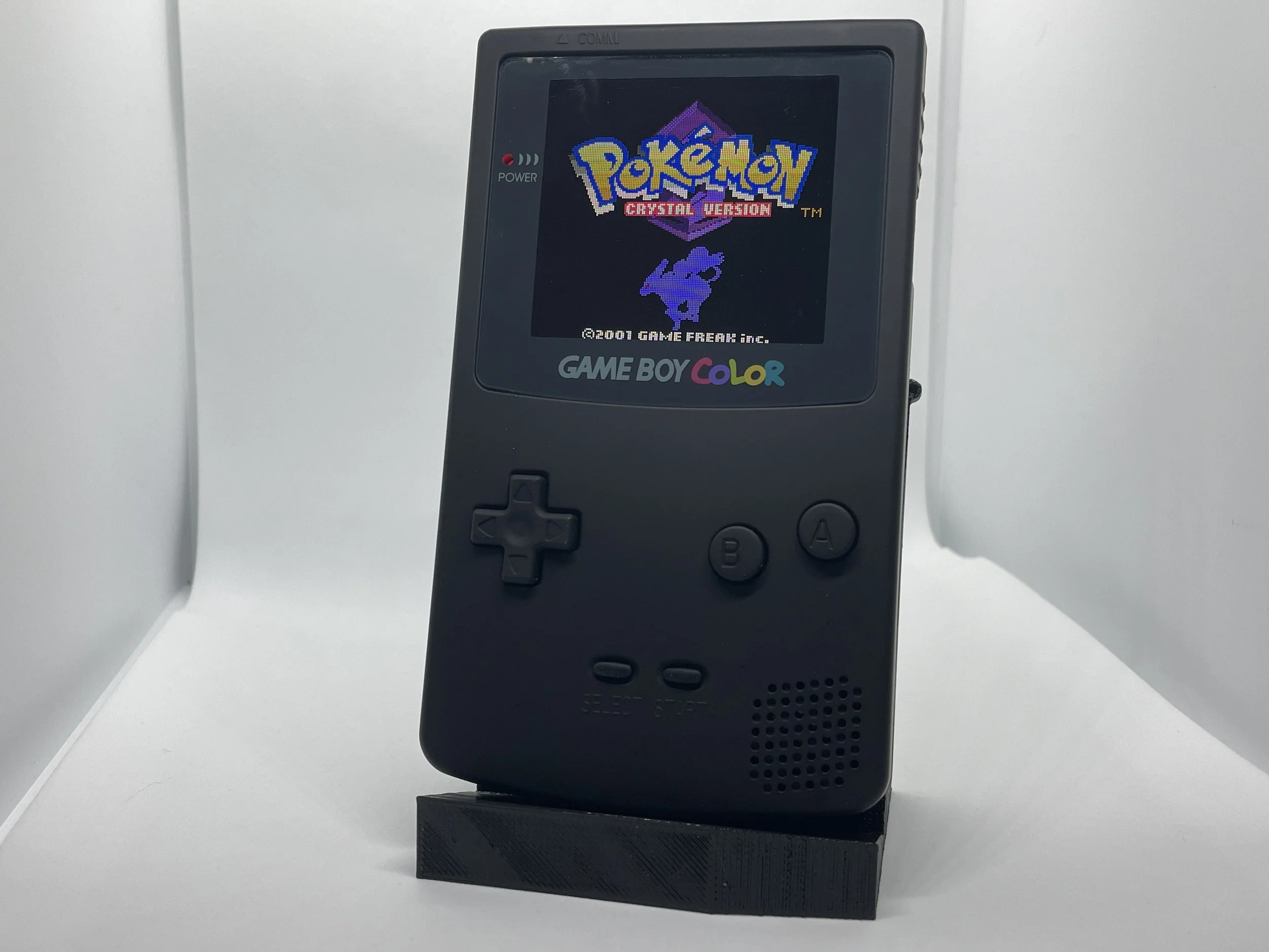 A black Game Boy Color handheld gaming console displaying the title screen of Pokémon Crystal Version, with a Pokémon sprite and the game logo.