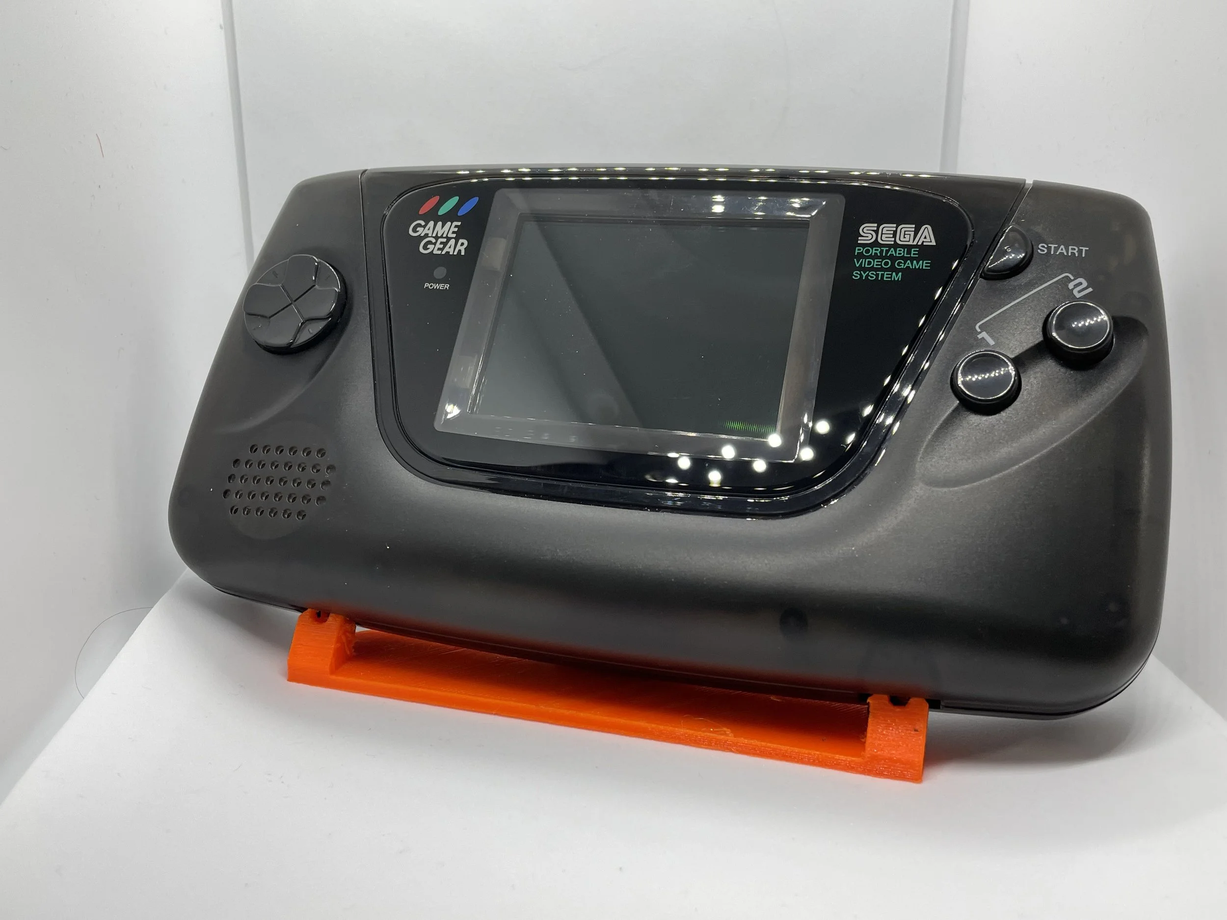 SEGA Game Gear portable video game system with black casing, a screen in the center, control buttons on the right, and a directional pad on the left, placed on an orange stand against a white surface.