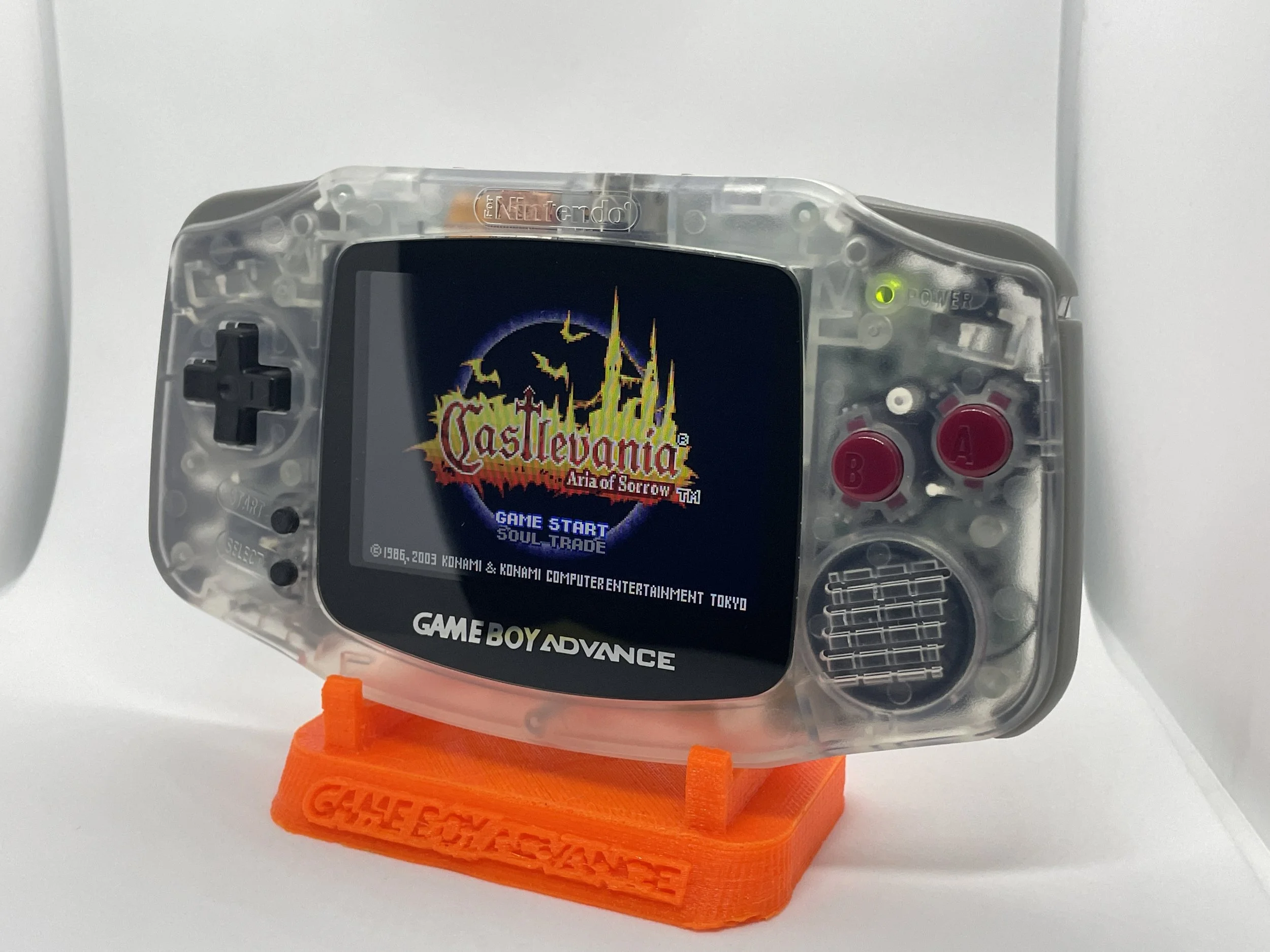 A clear, custom-printed Game Boy Advance with the game Castlevania: Aria of Sorrow displayed on its screen, mounted on an orange stand.