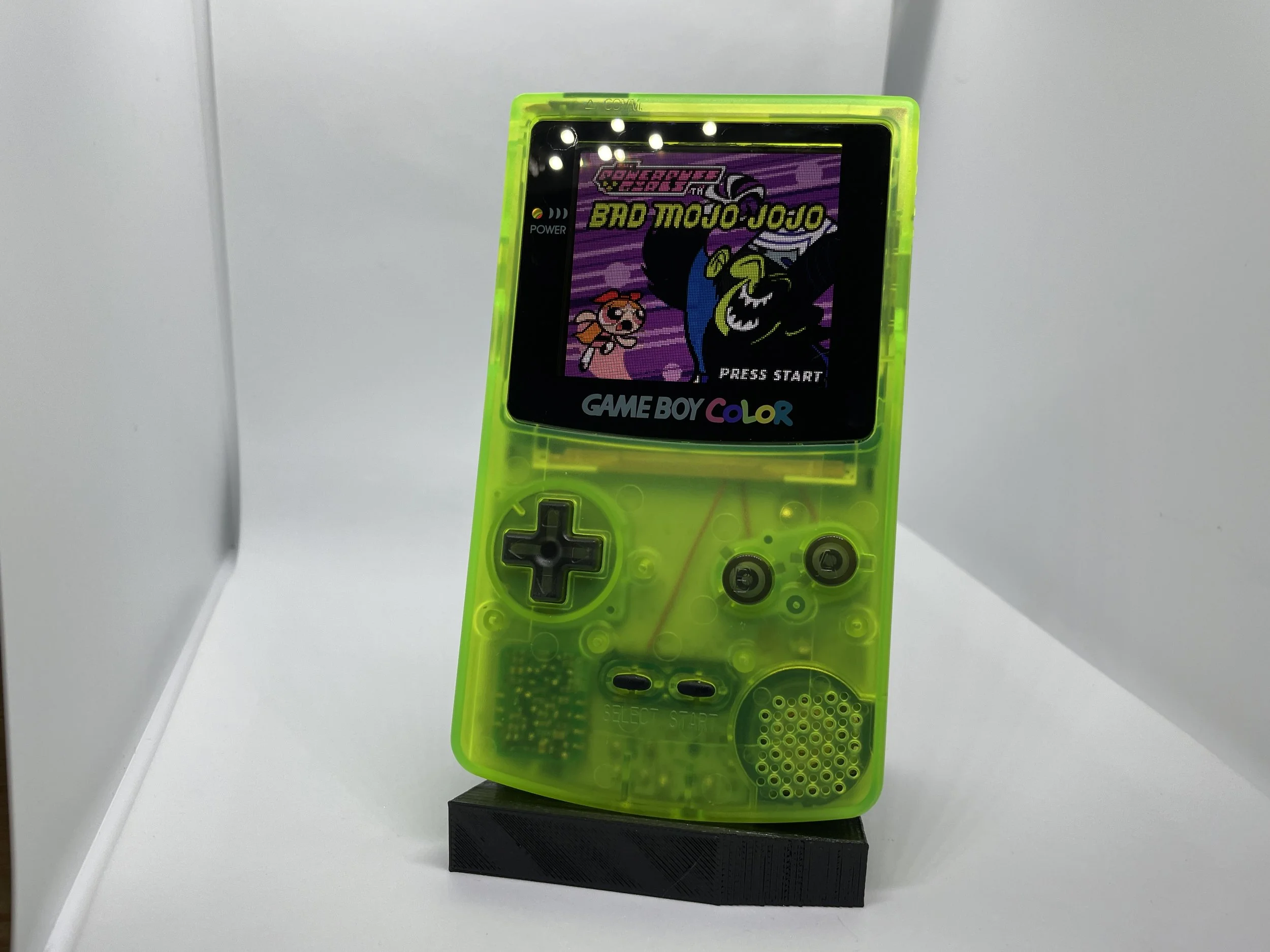 Neon green Game Boy Color handheld gaming console displaying the title screen of the game 'Crack Attack' with a cartoon character, a pink creature, and a moon in the background, placed on a black stand against a white background.