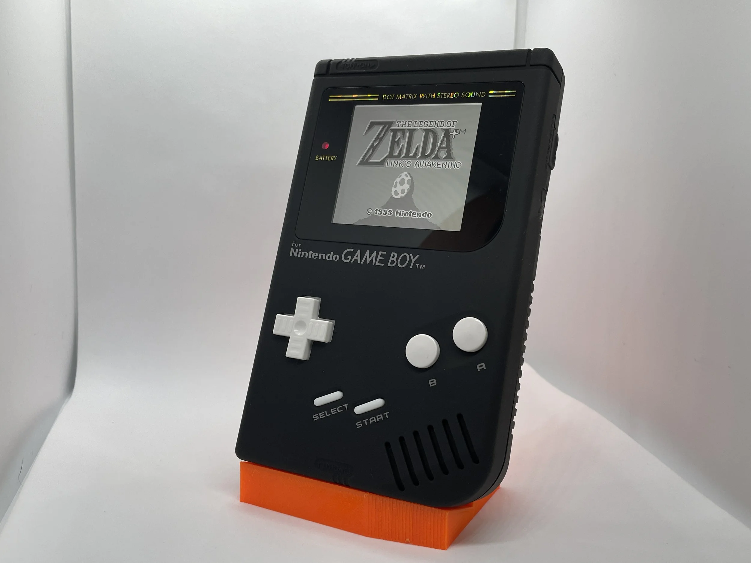 A vintage black Nintendo Game Boy handheld console displaying The Legend of Zelda: Link's Awakening game on its screen, placed on an orange stand against a white background.