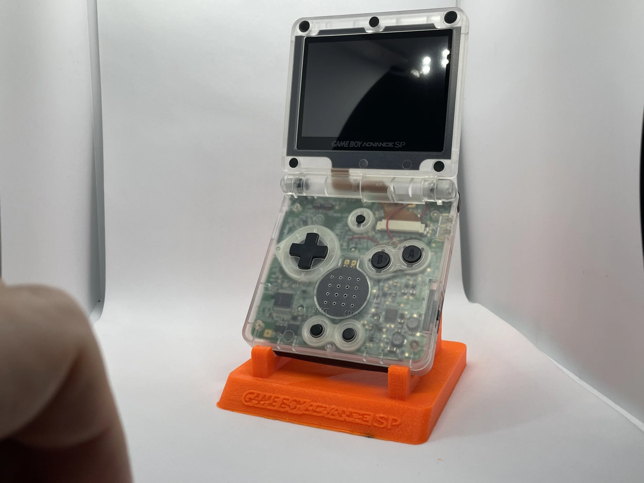 A transparent gaming handheld device with a visible circuit board, placed on an orange stand labeled 'GAME BOY ADVANCE SP', against a white background.