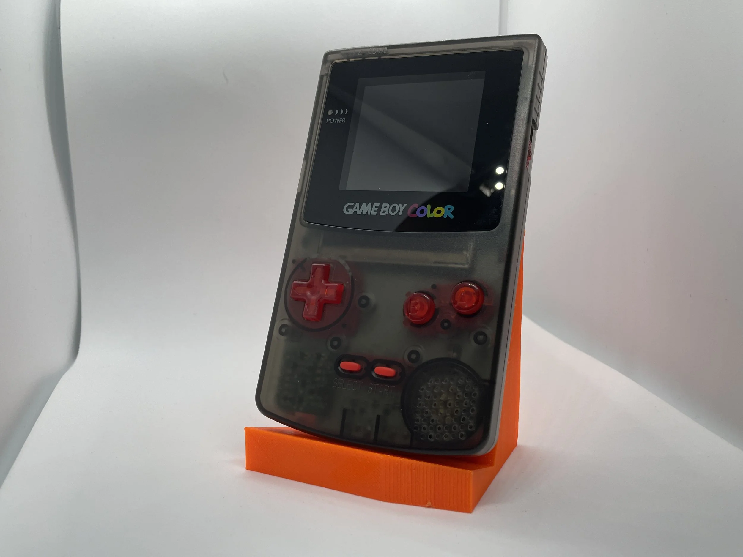 A vintage Game Boy Color handheld gaming device with a translucent gray body and red buttons, displayed on an orange stand against a white background.
