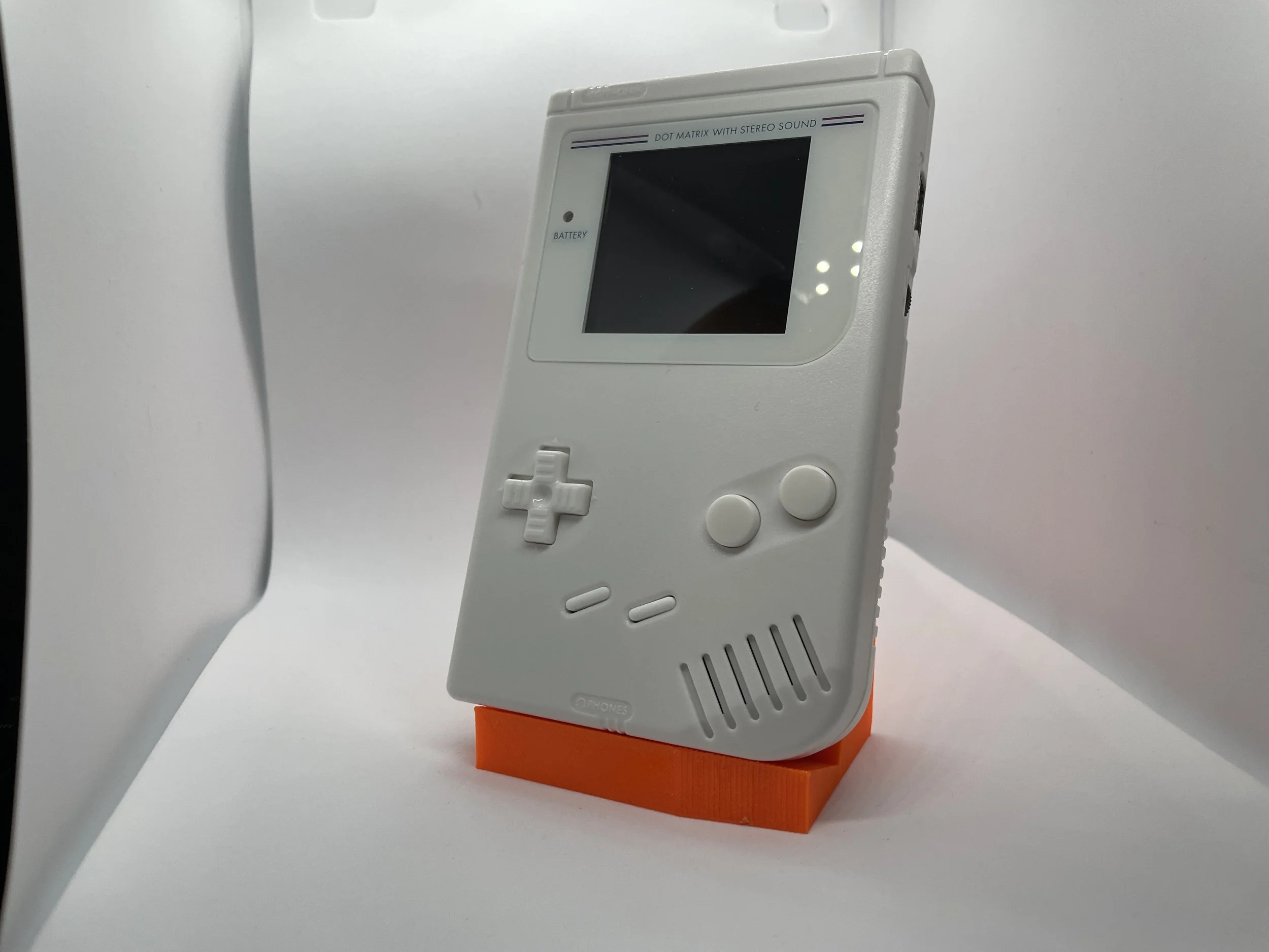 A white Nintendo Game Boy Classic with gray buttons, standing upright on an orange stand against a plain white background.