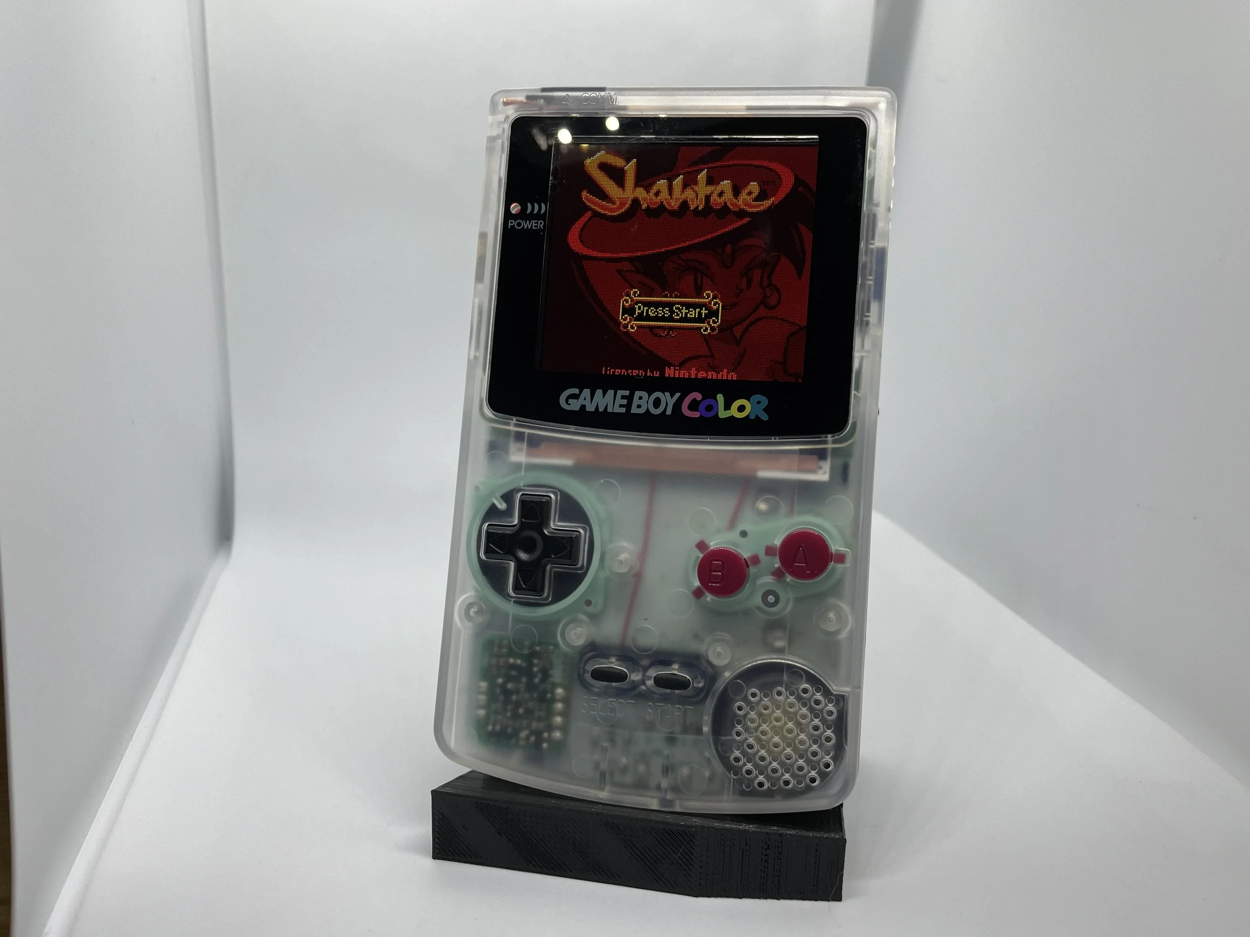 A transparent Game Boy Color with a custom design on the screen and pink A and B buttons, displaying the start screen of the game 'Shantae'.