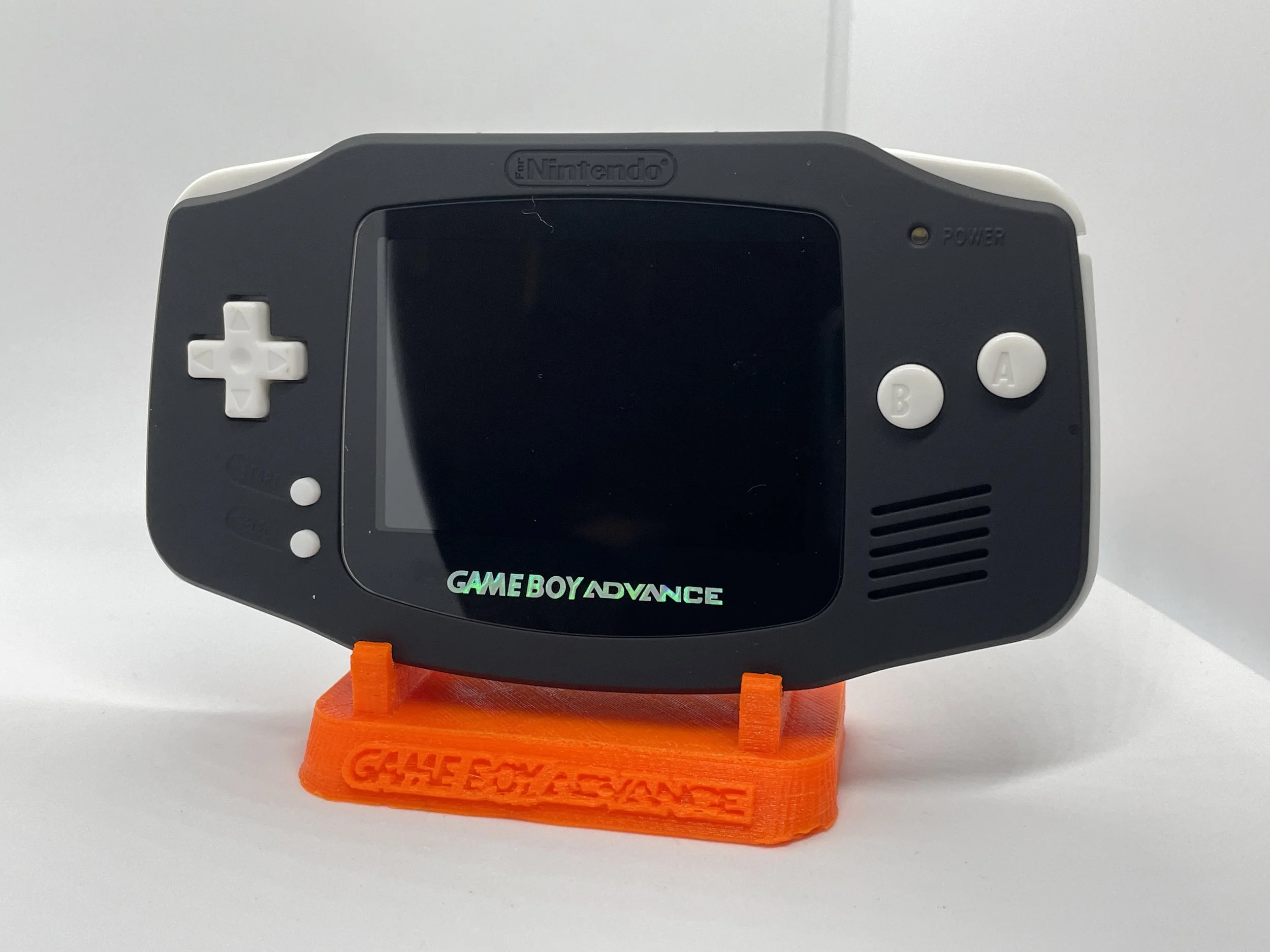 A Nintendo Game Boy Advance handheld gaming console on a bright orange stand that reads 'GAME ESCAPE' in red. The device is black with white buttons and a blank screen.