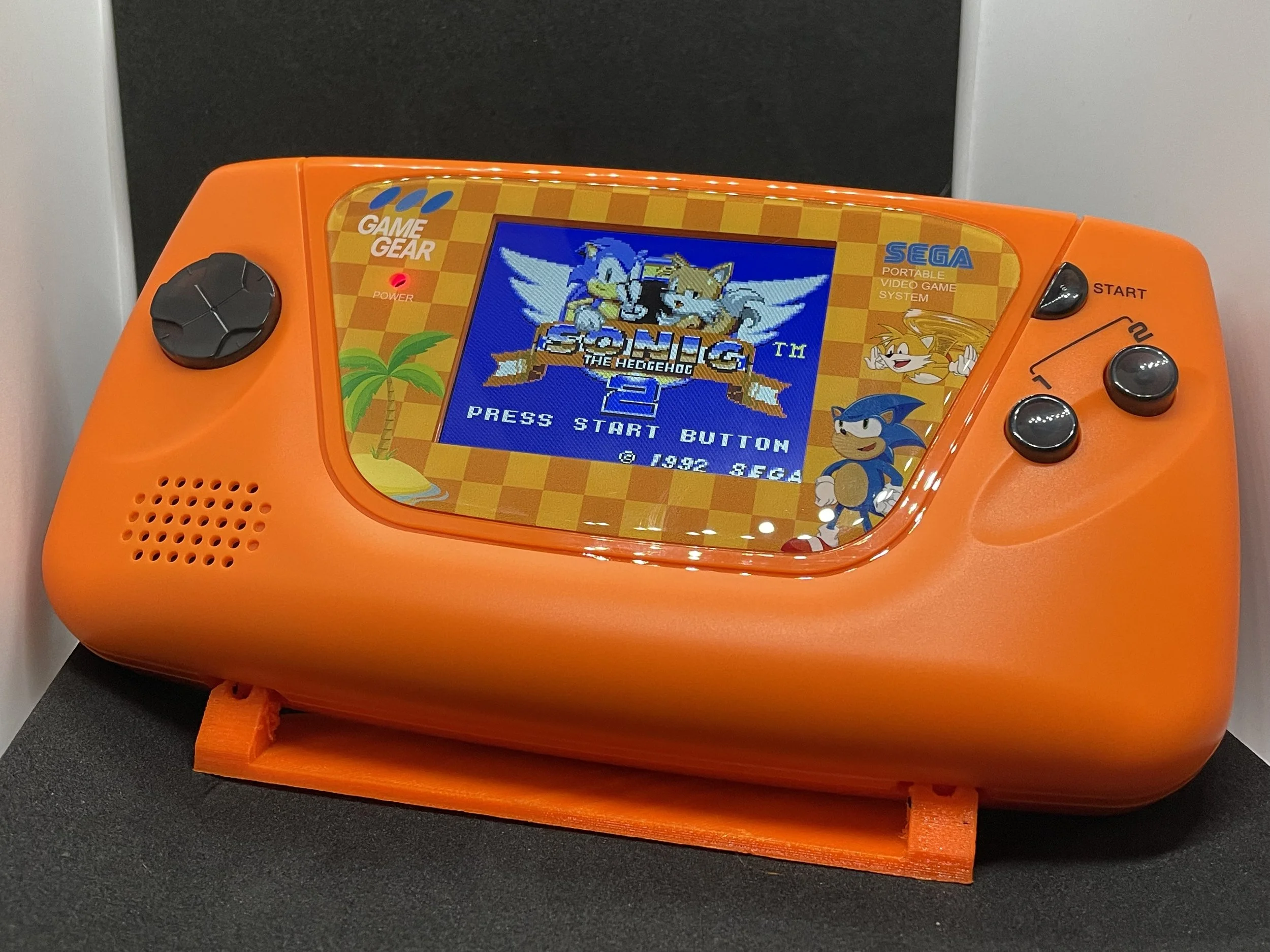 Orange handheld Sega game console with Sonic the Hedgehog game loaded on the screen, placed on a black surface.
