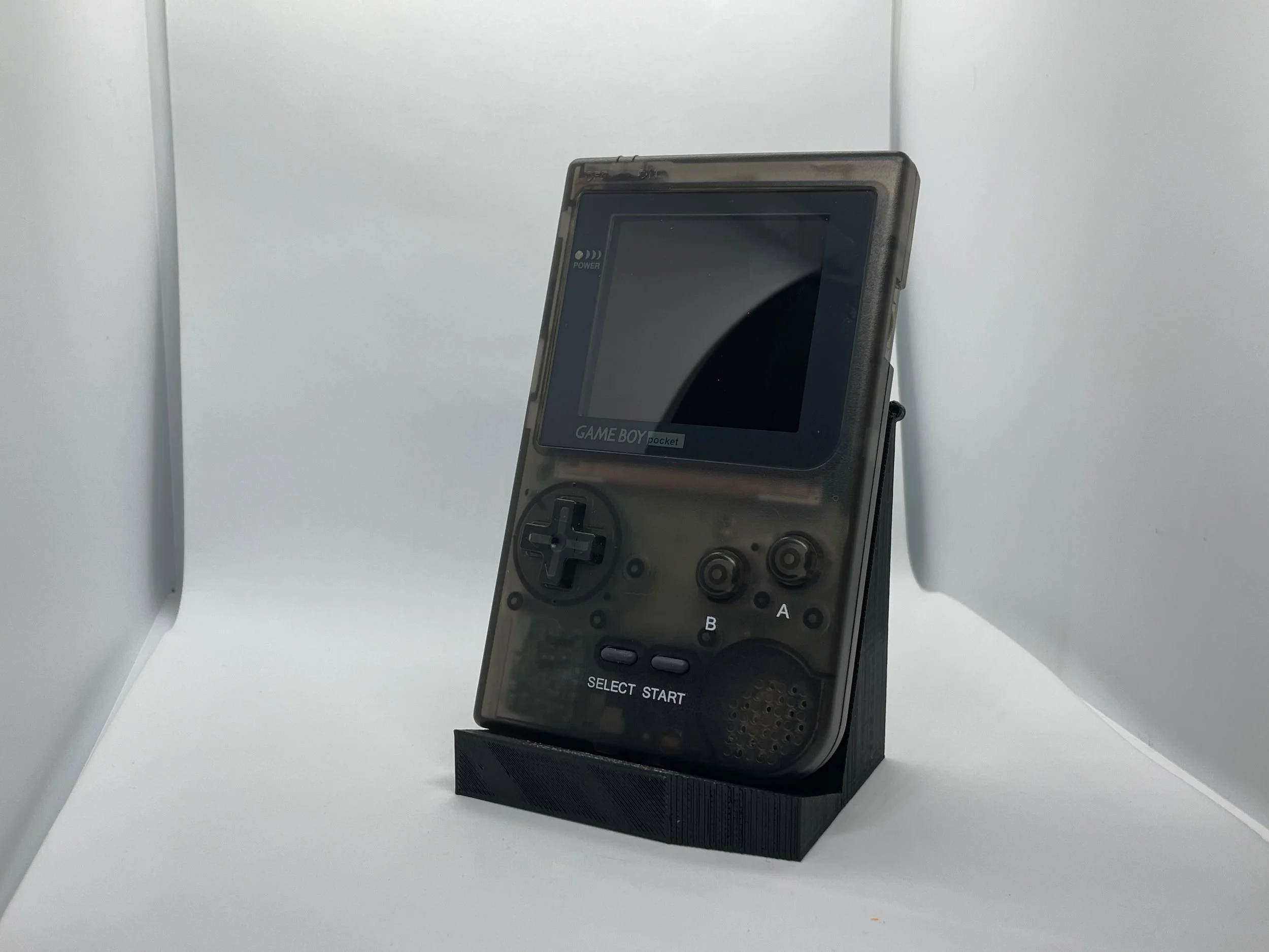 A clear plastic-clad Sony Game Boy Pocket handheld gaming device on a black stand, with a blank screen and gray buttons, set against a white background.