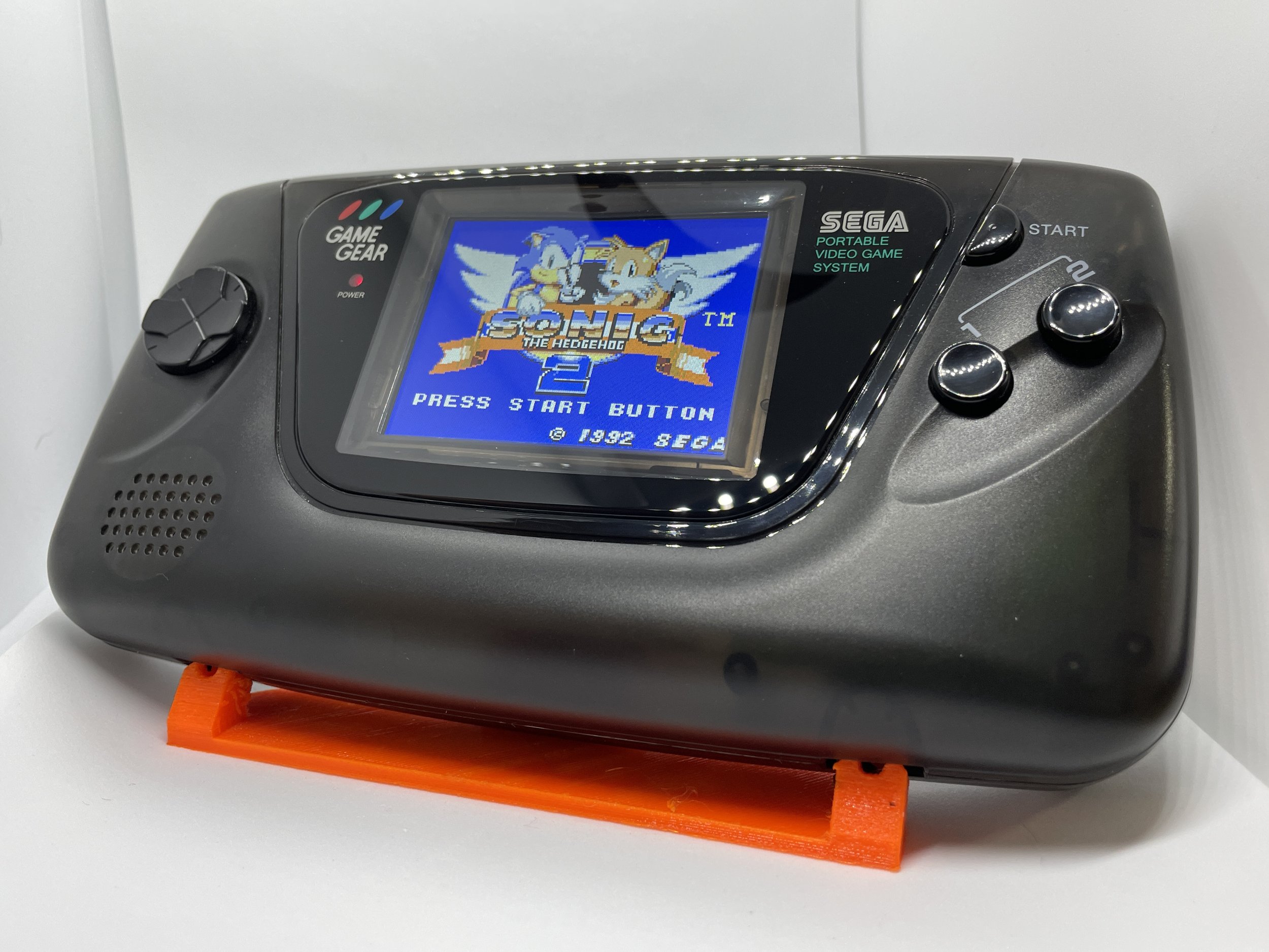 A portable Sega Game Gear gaming console displaying the title screen of Sonic the Hedgehog 2 with Sonic and Tails characters.