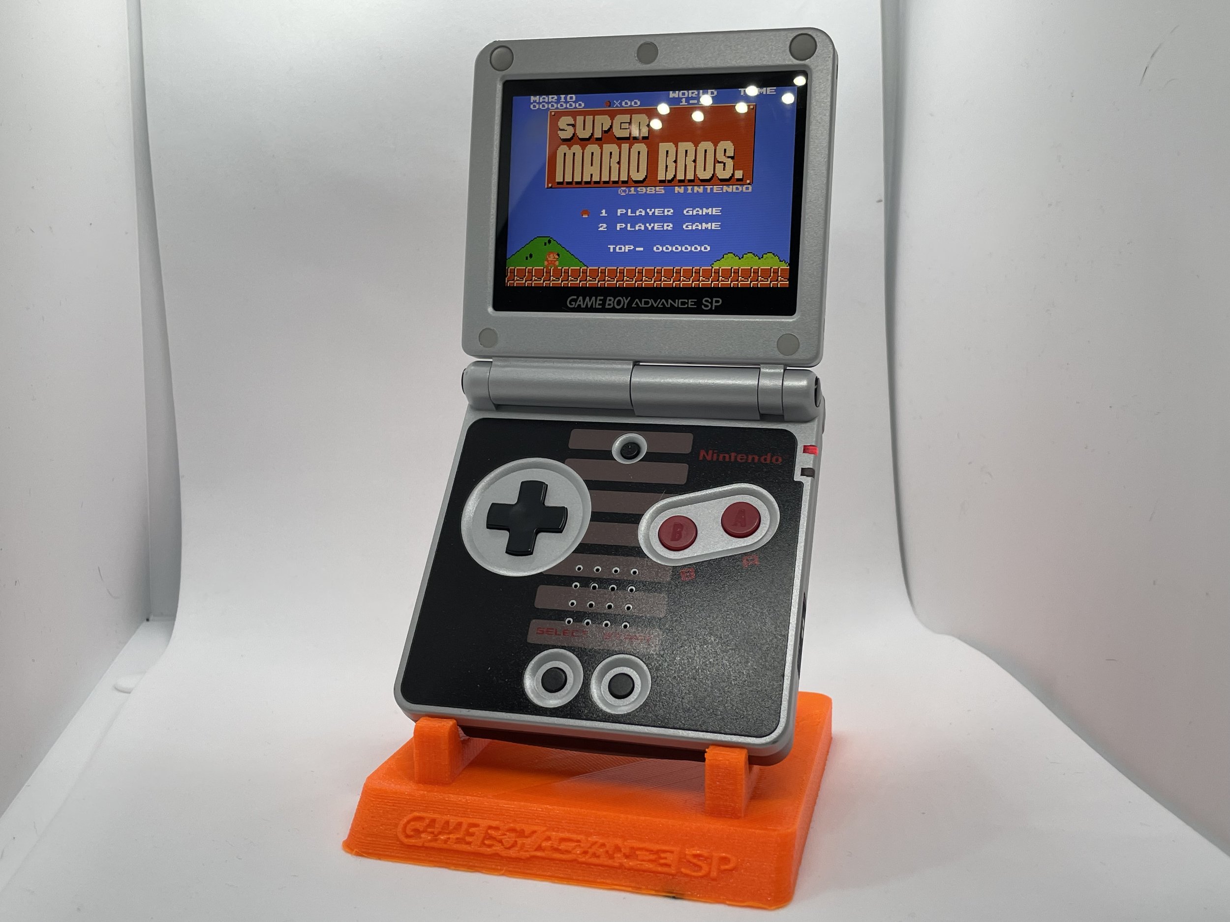 A mini replica of a Nintendo Game Boy Advance SP holding the game Super Mario Bros., with a white background and an orange stand labeled 'GAME BOY ADVANCE SP.'