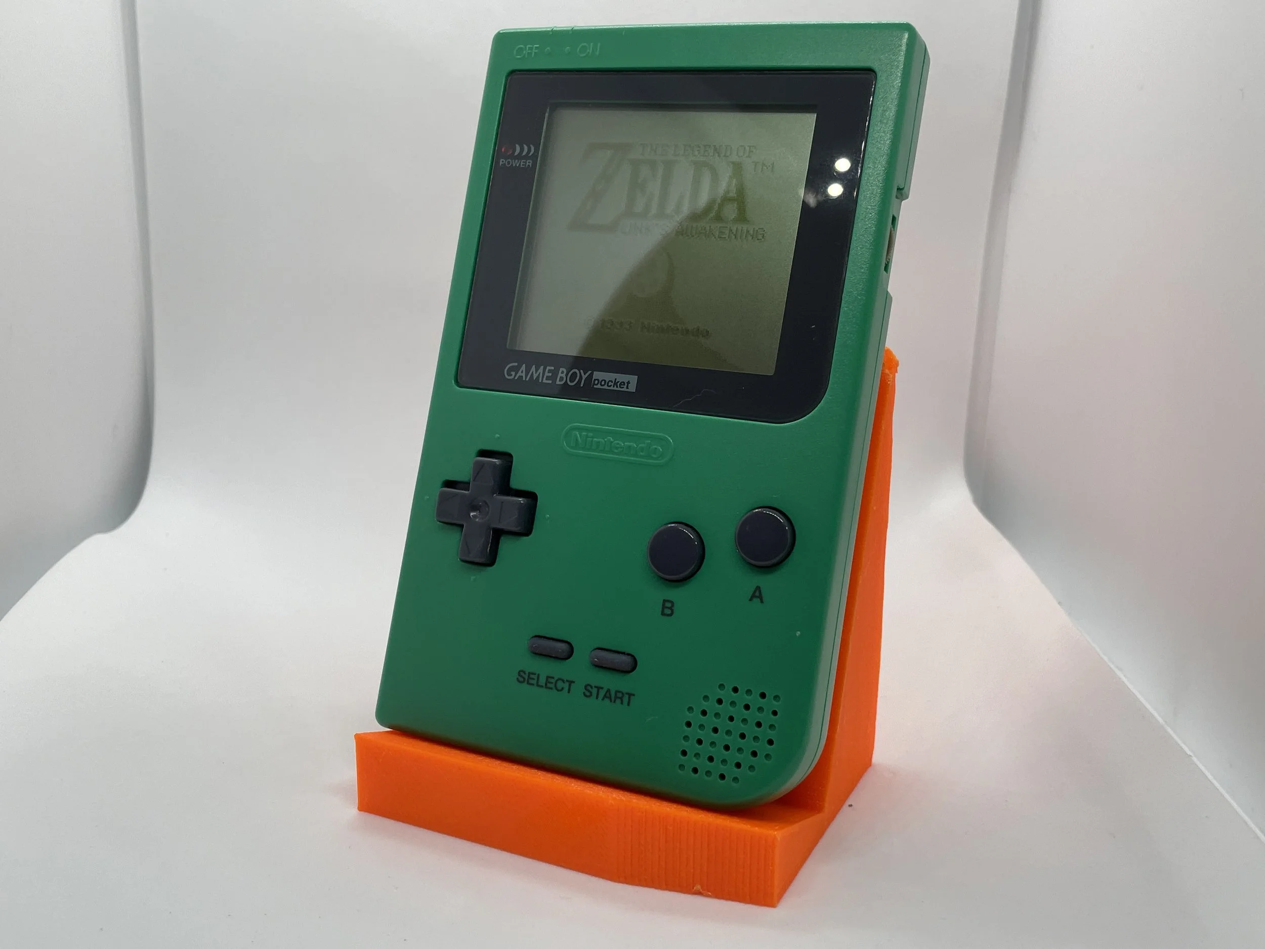 A green Nintendo Game Boy Pocket placed on an orange stand, displaying the game "The Legend of Zelda: A Link to the Past & Four Swords" on its screen.