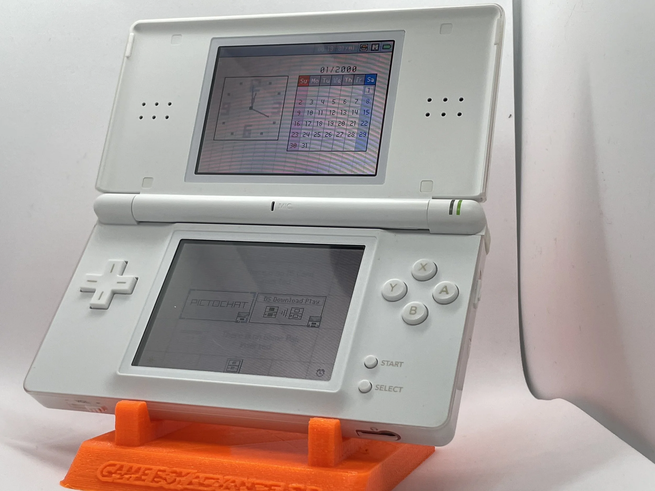 A white Nintendo DS handheld gaming console with dual screens, placed on an orange stand. The top screen displays a calendar for January 2000, and the bottom screen shows a menu with app icons.