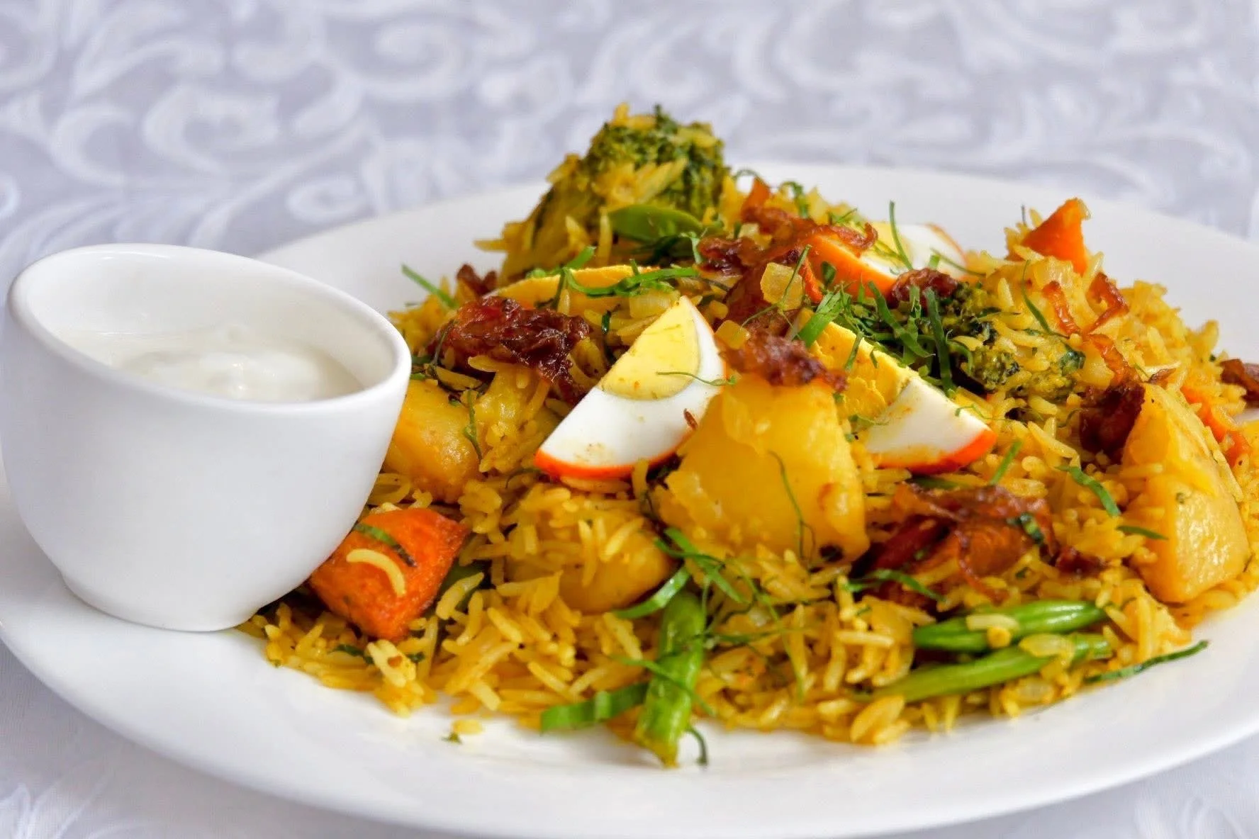 Plate of yellow rice mixed with vegetables, garnished with chopped herbs, and topped with slices of boiled egg, served with a small container of creamy white sauce on the side.