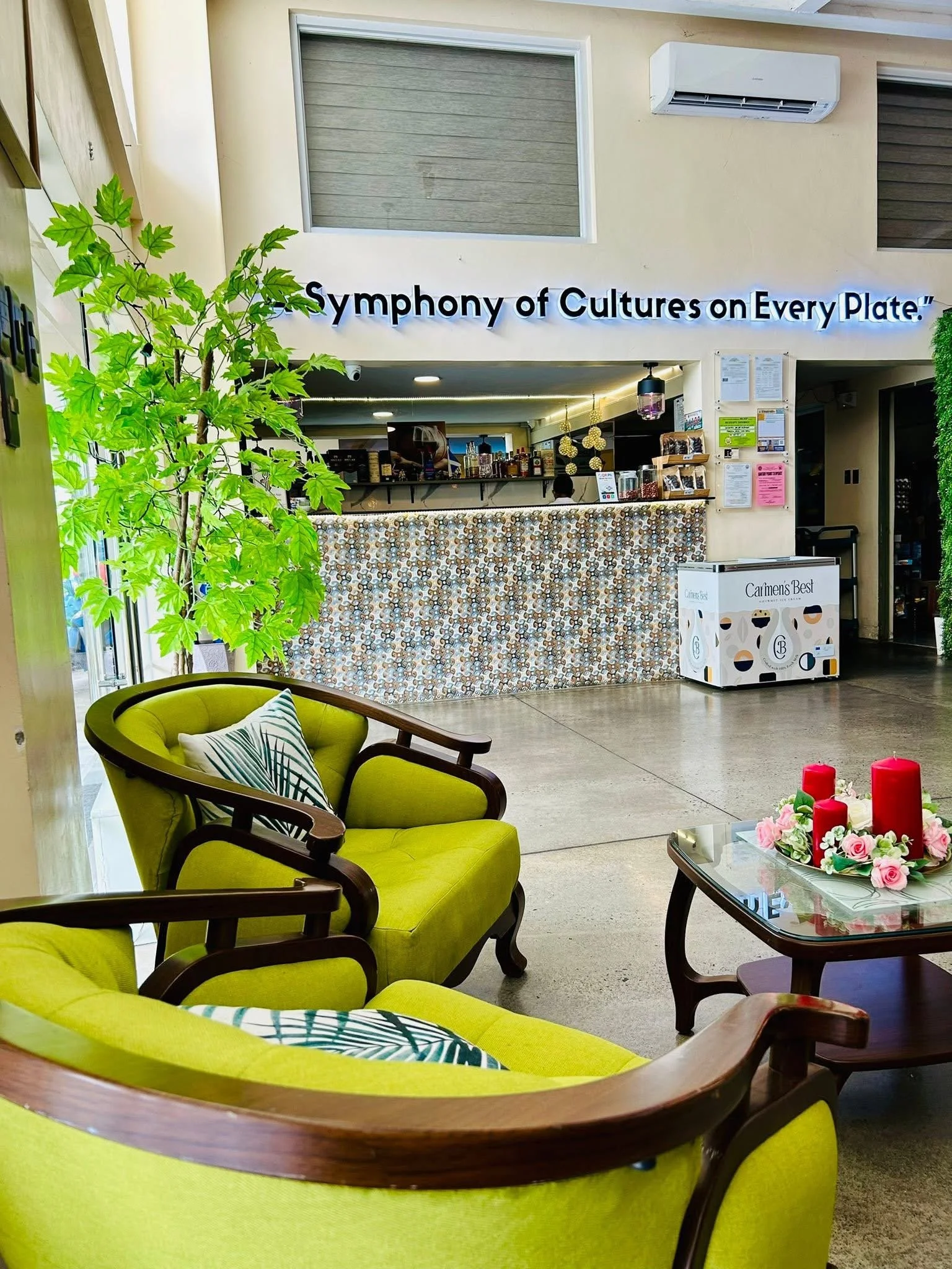 Interior of a cafe or restaurant with a green cushioned chair in the foreground, a floral arrangement with candles on a glass table, a counter area with coffee and items behind it, a green leafy plant on the left, and a sign that reads 'Symphony of Cultures on Every Plate' hanging above the counter.