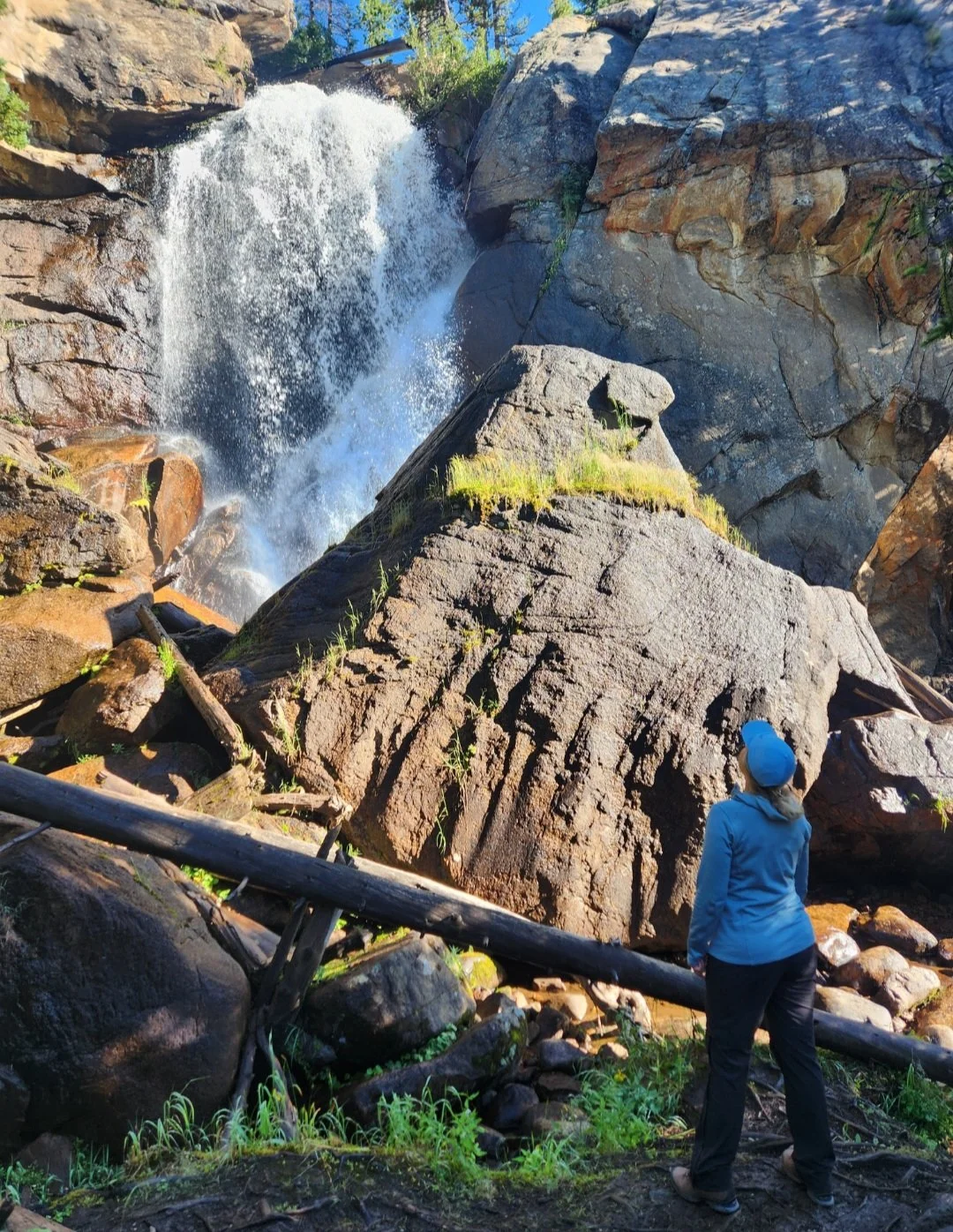 A person in a blue jacket and blue hat standing in front of a large boulder near a waterfall in a rocky outdoor setting looking up at the 40-foot Ouzel Falls on a women-only Ouzel Falls women-only guided hiking tour with Femme Trek.