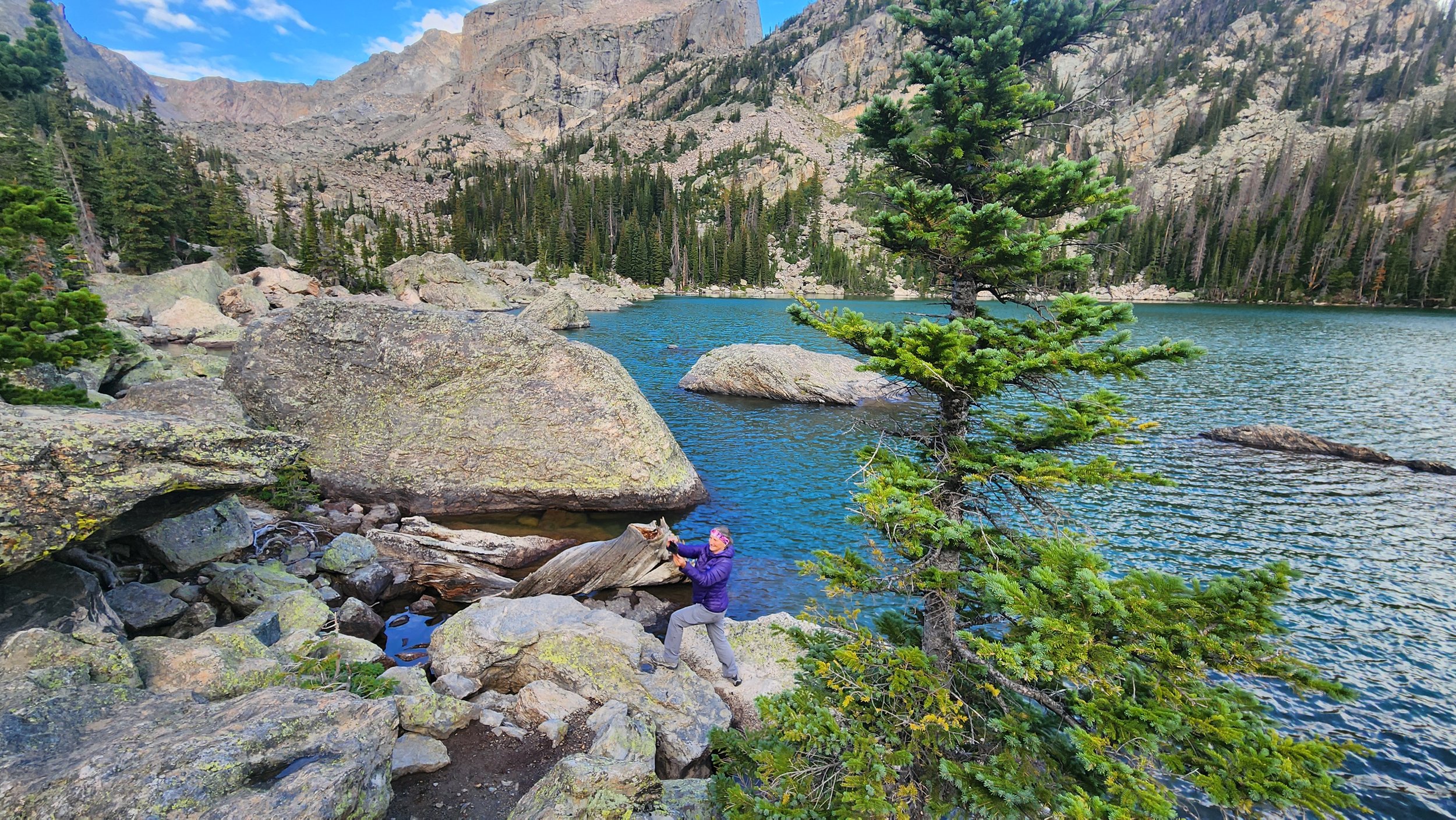 A woman in a purple jacket and gray pants taking a photo by a lake surrounded by rocks, pine trees, and mountains under a partly cloudy sky while on a women-only guided hiking tour to Lake Haiyaha in Rockly Mountain National Park with Femme Trek.