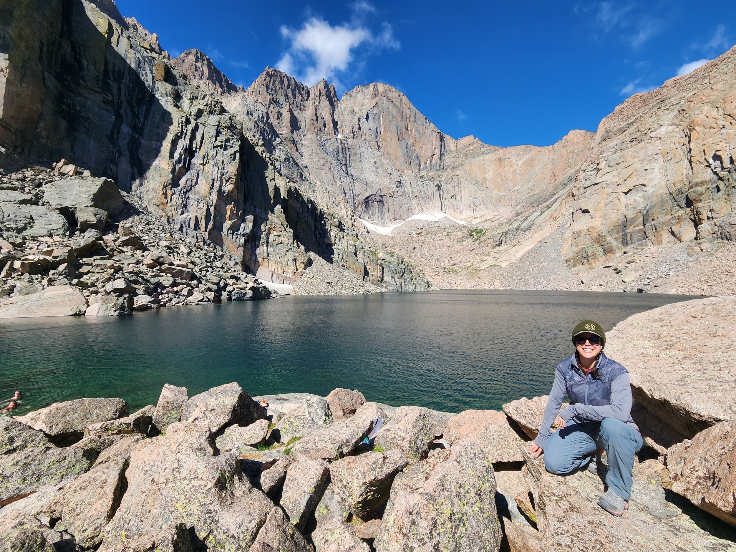 Femme Trek guest kneels down on granite rocks in front of Chasm Lake with Longs Peak in the background on a women only guided hiking tour with Femme Trek in Rocky Mountain National Park