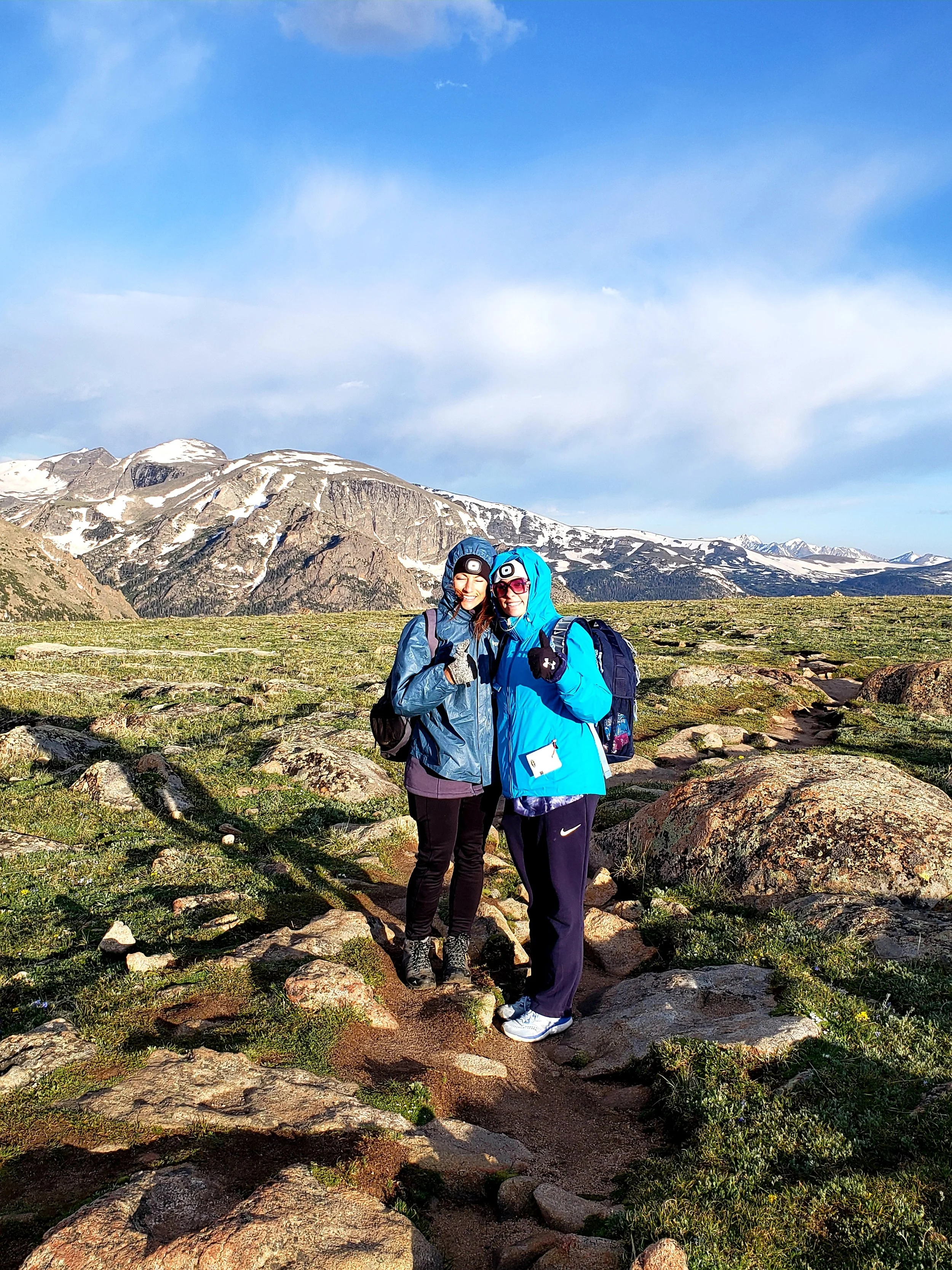 Two women in outdoor gear standing close together on a rocky trail with mountain ranges and partly cloudy sky in the background. Photo by Femme Trek on a women's private guided hiking tour with Femme Trek in Rocky Mountain National Park.